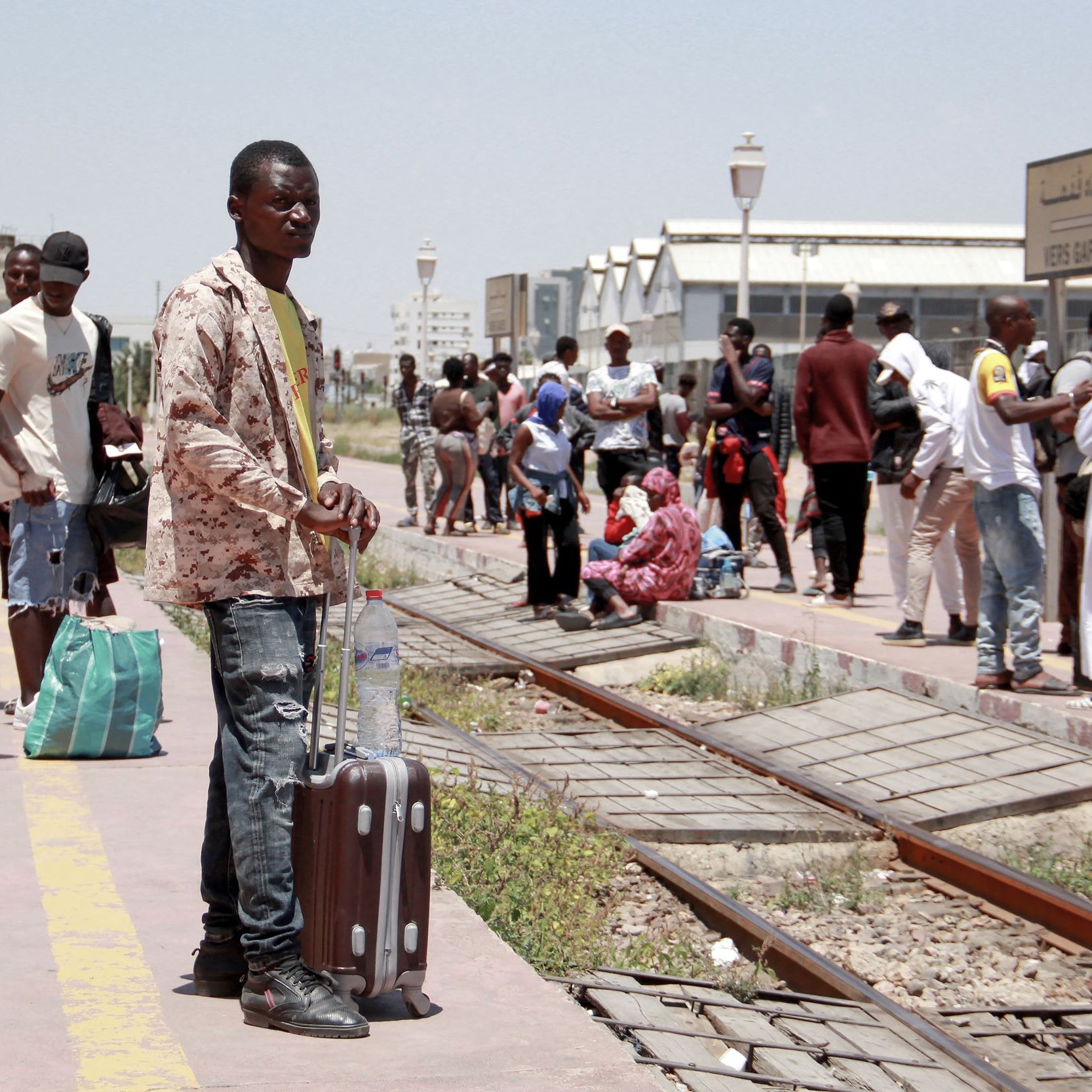 L'image montre une station de train animée. Un homme debout, tenant une valise, semble attendre. En arrière-plan, on peut voir d'autres personnes : certaines attendent assises, d'autres se déplacent avec des sacs. L'environnement est ensoleillé, et il y a des bâtiments visibles au loin. La scène évoque un moment de déplacement et d'attente.