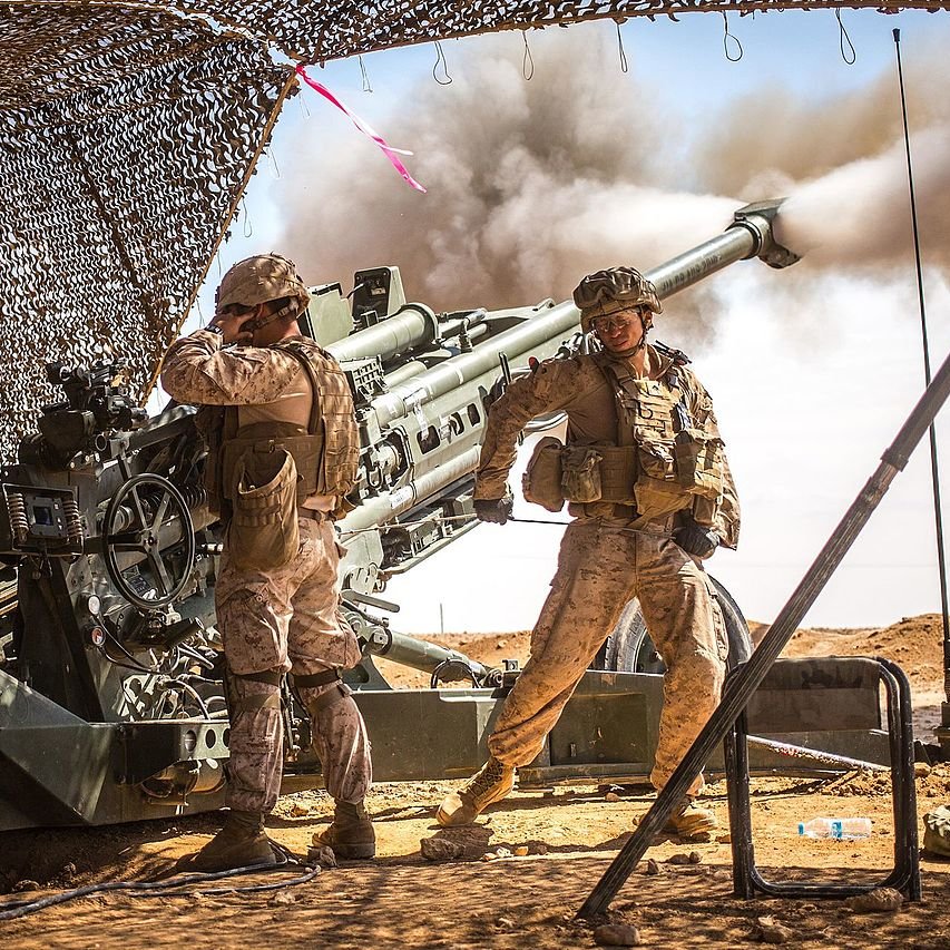 L'image montre deux soldats en uniforme militaire, probablement dans un environnement désertique. Ils sont proches d'une grande pièce d'artillerie qui est en train de tirer, créant un nuage de fumée. L'un des soldats semble gérer l'équipement tandis que l'autre se tient debout, surveillant la situation. Un camouflage est utilisé pour dissimuler l'équipement militaire, et le paysage environnant est aride avec un ciel dégagé. L'atmosphère semble tendue et concentrée, rappelant des opérations militaires.