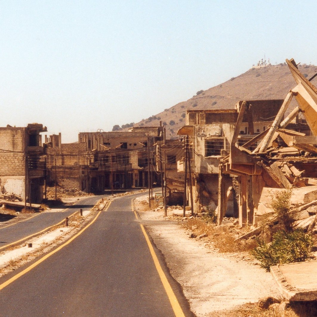 L'image montre une scène de destruction et de désolation. On peut voir une route déserte, bordée de bâtiments en ruines et de débris. Les structures semblent fortement endommagées, avec des murs effondrés et des fondations exposées. À l'arrière-plan, une colline se dresse, mais l'accent est principalement mis sur la destruction des habitations. L'atmosphère est empreinte de tristesse et d'abandon, suggérant que cet endroit a subi des conflits ou des catastrophes.