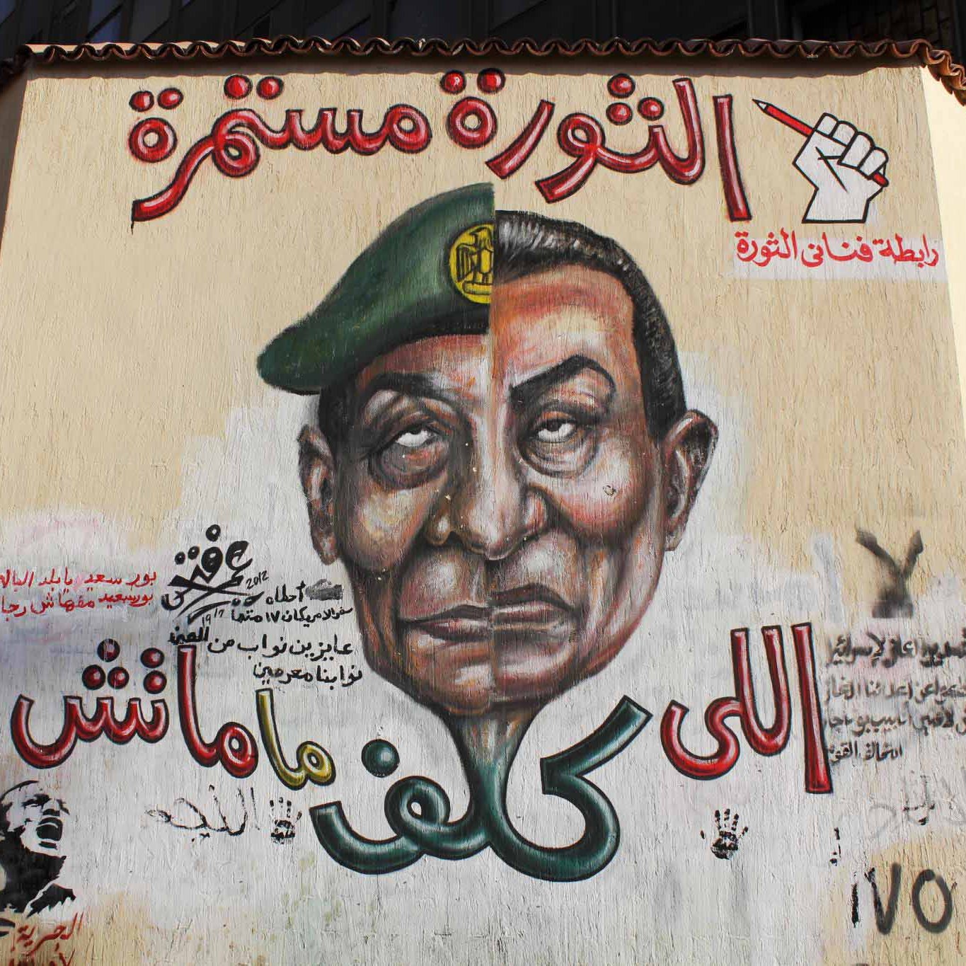 L'image montre une fresque murale sur un mur, représentant un homme avec un uniforme militaire et un képi. Son expression est sérieuse et emblématique. Autour de cette image, on trouve des inscriptions en arabe, dont certaines font allusion à une continuité de la révolution. Le style artistique est vibrant et engagé, évoquant des thèmes de soulèvement et de résistance. Des symboles de révolte ou de mouvement peuvent également être présents, soulignant un message politique fort.