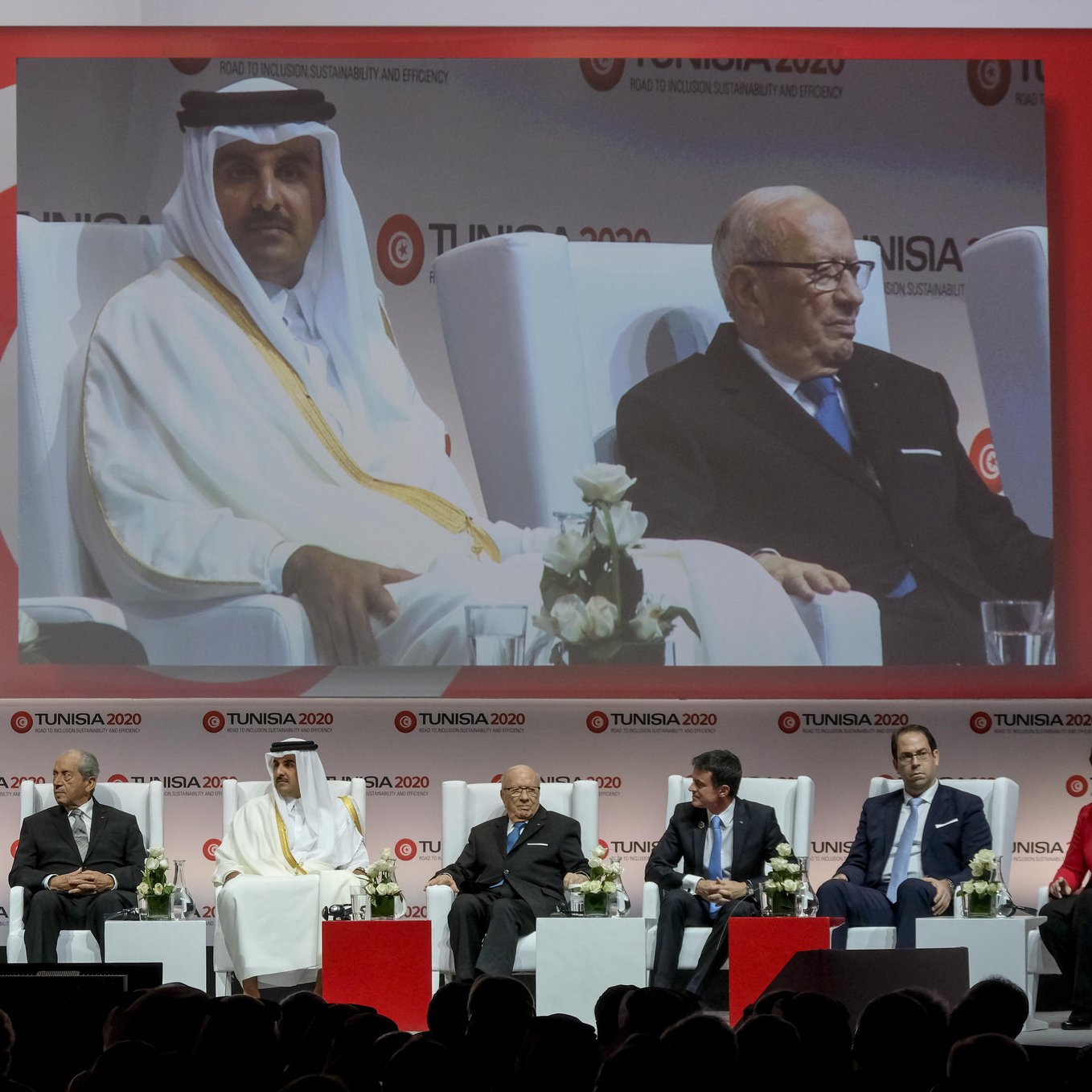 L'image montre une scène de conférence ou de séminaire avec plusieurs intervenants assis sur des chaises, sur une scène décorée avec des fleurs blanches. Au centre, il y a un grand écran affichant un homme en costume, vraisemblablement un orateur ou un participant à l'événement. Les participants semblent représentés sur une plateforme, prenant part à une discussion ou à une cérémonie. Le fond est décoré avec des motifs et des logos liés à l'événement "TUNISIA 2020". L'ambiance semble professionnelle et formelle.