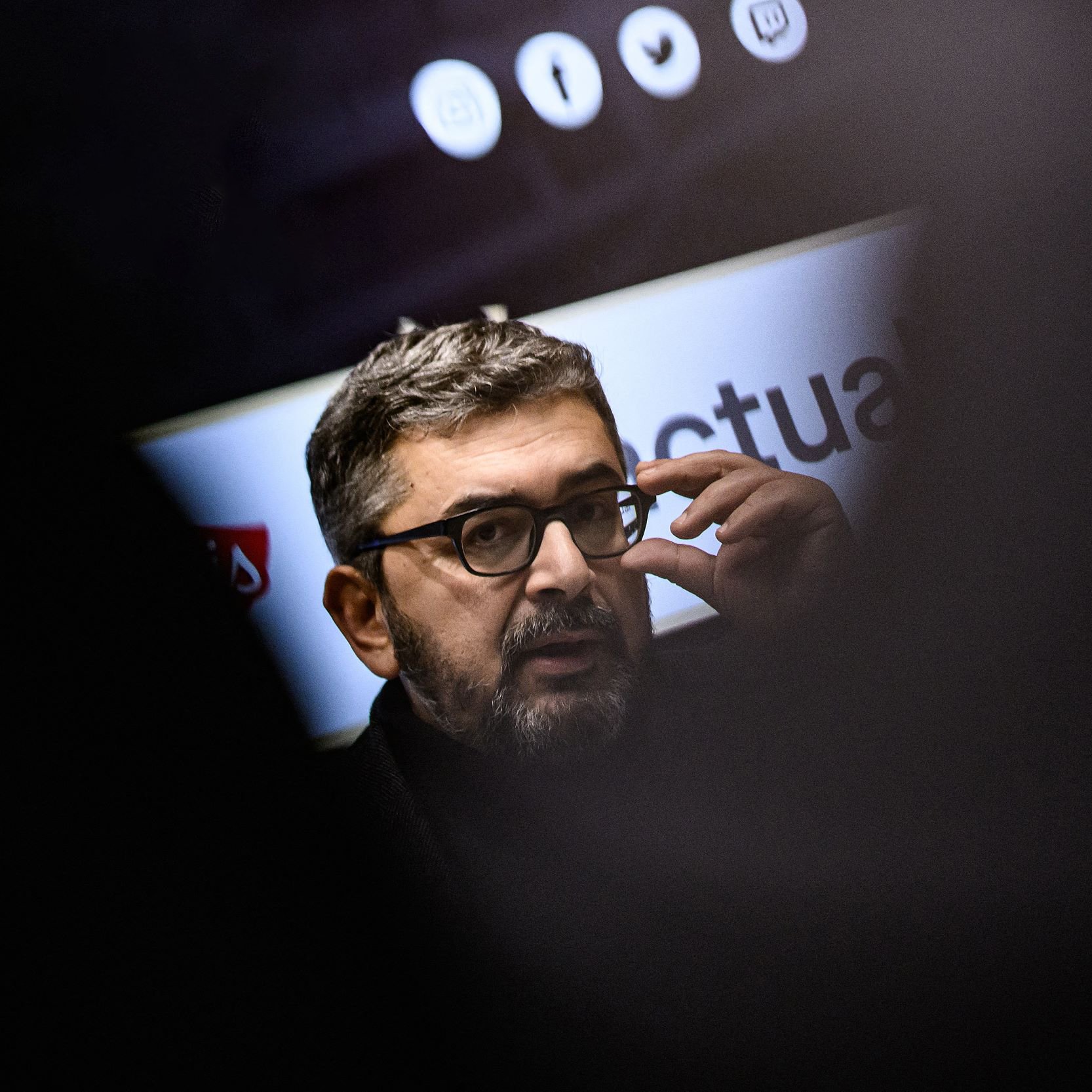 L'image montre un homme portant des lunettes et ayant une barbe courte. Il semble engagé dans une conversation ou une présentation, en touchant son visage avec une main. En arrière-plan, des éléments graphiques ou des logos sont flous, ce qui suggère qu'il se trouve dans un cadre professionnel ou public. L'éclairage met en avant son visage, tandis que des silhouettes floues d'autres personnes sont visibles autour.