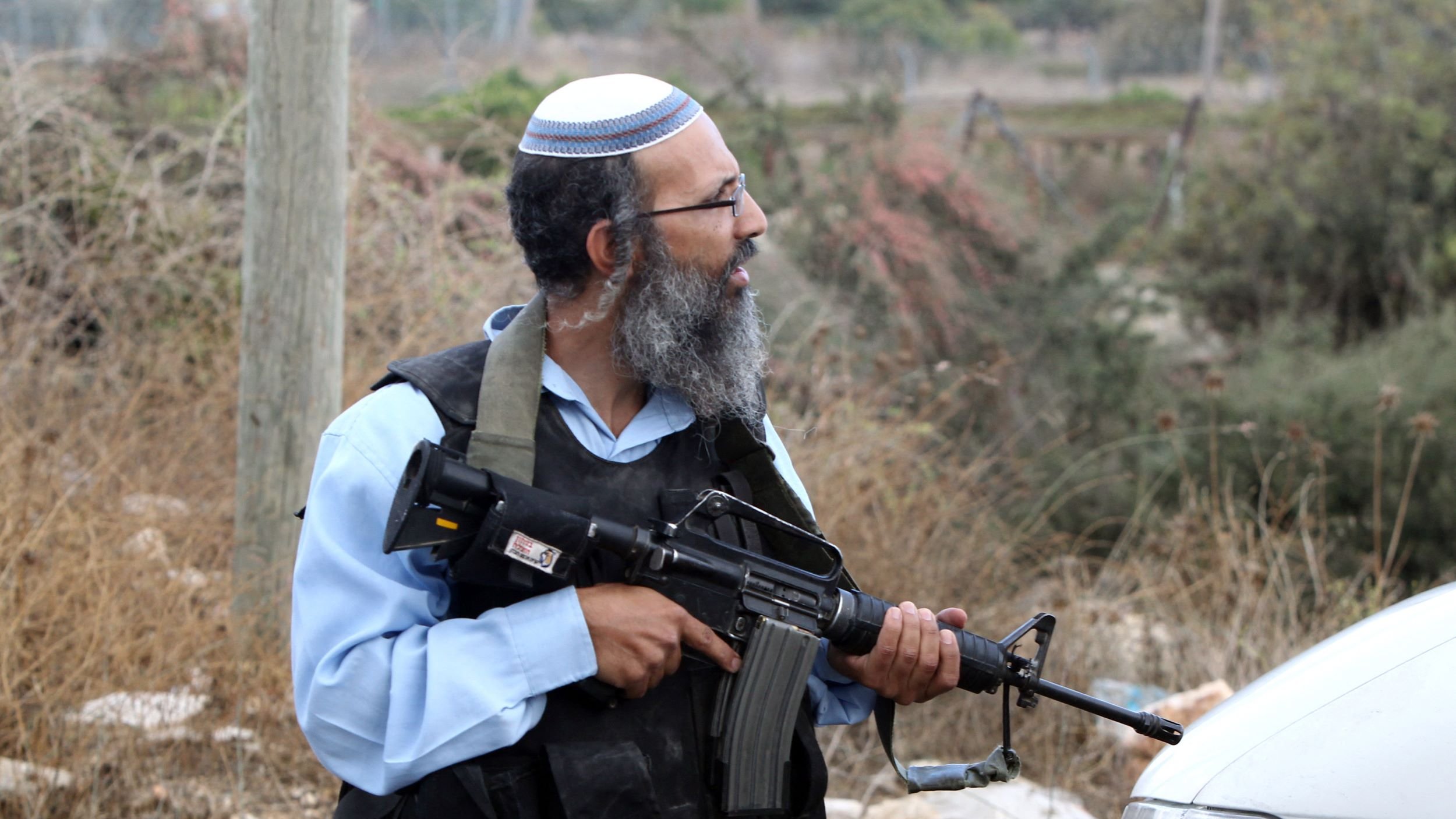 L'immagine mostra un uomo in piedi in un ambiente all'aperto con vegetazione secca sullo sfondo. Indossa un kippah e un giubbotto tattico, e tiene in mano un fucile. L'atteggiamento e l'abbigliamento suggeriscono un contesto di sicurezza o di conflitto. Accanto a lui si vede un veicolo. La scena sembra essere in una zona rurale o periferica.