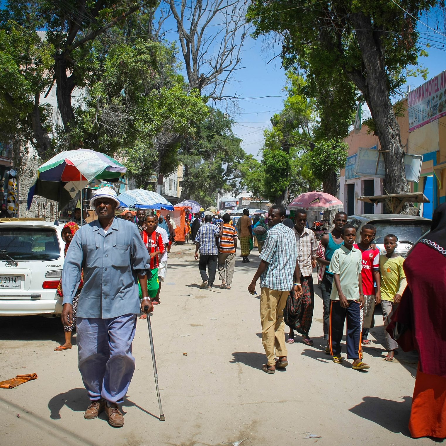 The image depicts a bustling street scene, likely in an urban area. There are several people walking along the road, some carrying umbrellas for shade. A few vehicles are parked along the sides, including cars that are mostly white. The atmosphere appears lively, with a variety of pedestrians, including a man using a walking cane. The buildings on either side are relatively simple, suggesting a community environment. Trees provide some greenery, enhancing the street's appearance. Overall, it captures a moment of daily life in a vibrant neighborhood.