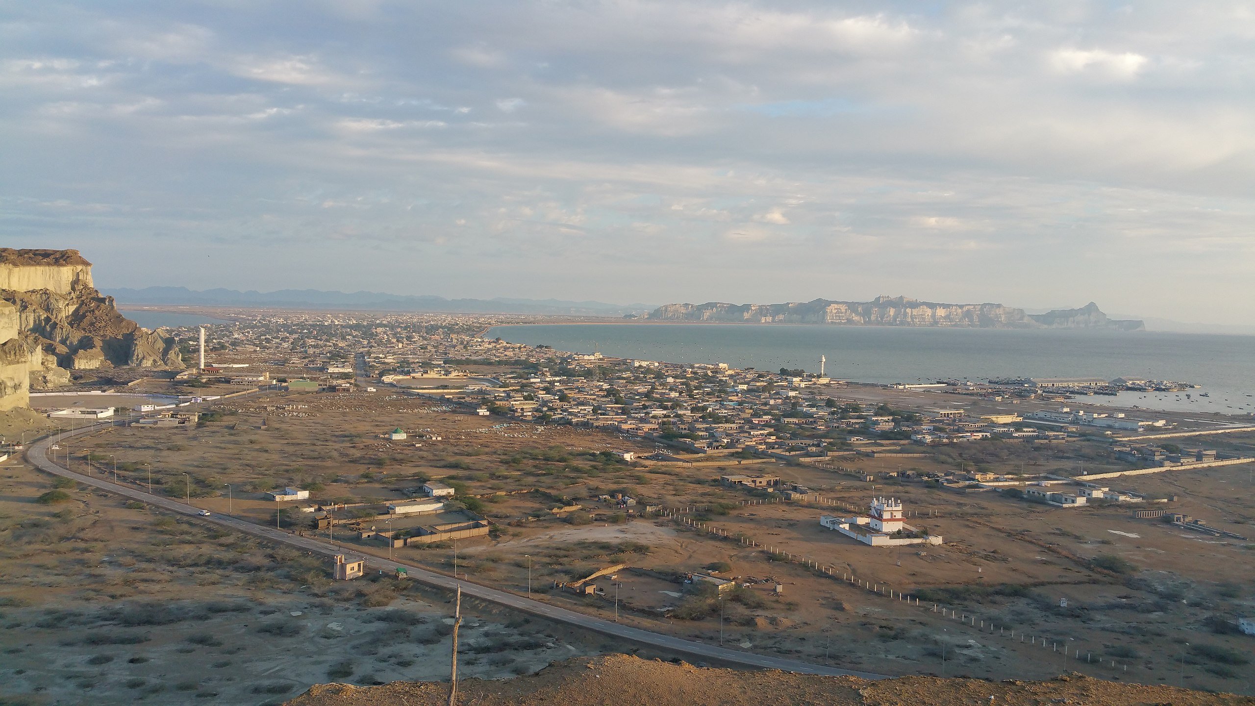 The image depicts a coastal landscape with a view overlooking a small town. In the foreground, there is a sparsely populated area with some buildings and roads, while the background features a scenic stretch of coastline meeting the sea. The sky is partly cloudy, casting soft light over the landscape. Distant mountains or cliffs are visible along the horizon, adding depth to the view. The overall atmosphere suggests a quiet, serene environment by the water.