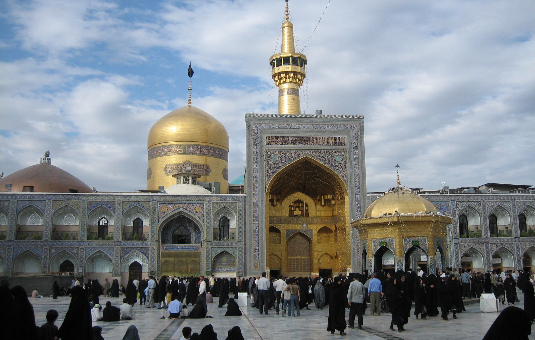 L'image montre un complexe religieux aux caractéristiques architecturales majestueuses. On peut y voir de grandes dômes, dont l'un est doré, qui attire particulièrement l'attention. Le bâtiment principal présente une grande porte ornée et des murs richement décorés. De nombreuses personnes, probablement des pèlerins, se rassemblent dans la cour, vêtues de vêtements traditionnels sombres. Le ciel est partiellement nuageux, ajoutant une ambiance spirituelle à l'ensemble du paysage.