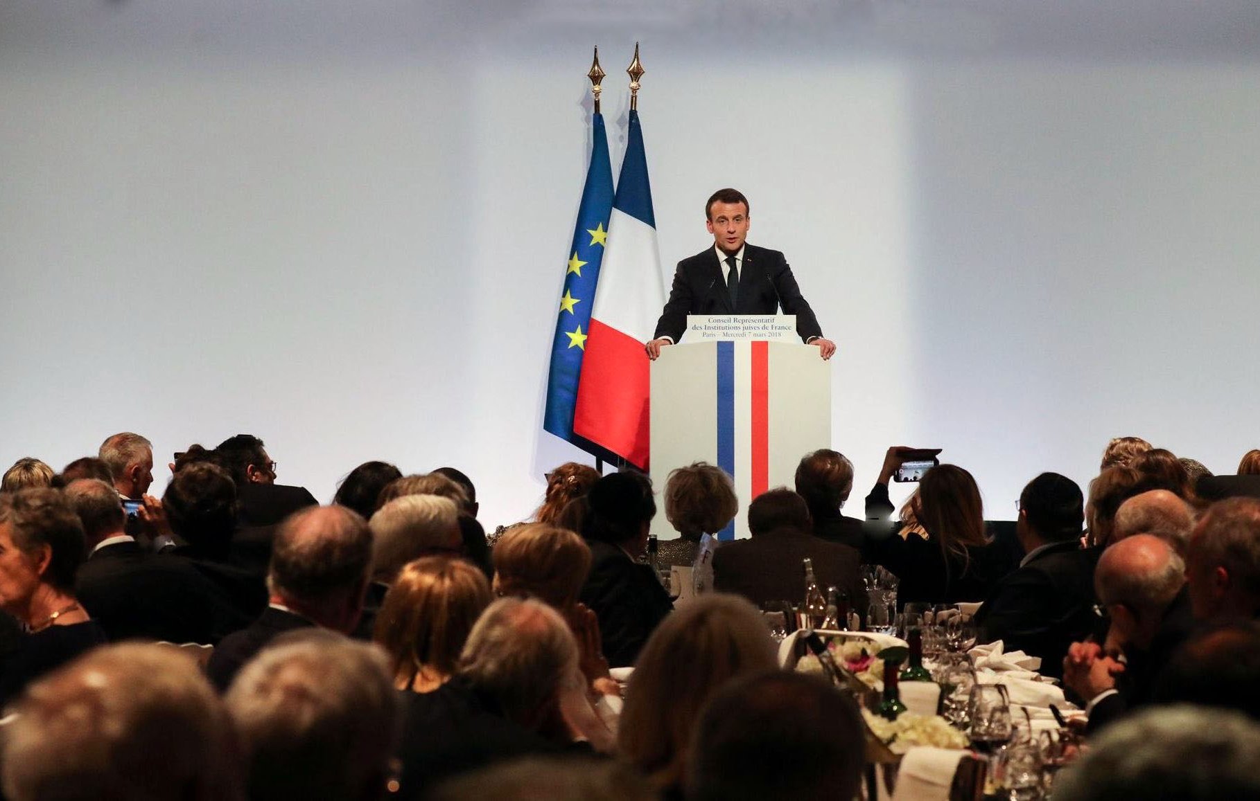 L'image montre un événement formel où une personne, probablement un homme politique, se tient devant un podium. Il parle à un public assis à des tables bien dressées, avec des couverts et des verres. En arrière-plan, on peut voir des drapeaux tricolores, bleu, blanc et rouge, représentant la France. L'atmosphère semble être celle d'un dîner officiel ou d'une réception. Les invités sont attentifs et nombreux, reflétant un moment de discours ou de présentation dans un cadre élégant.