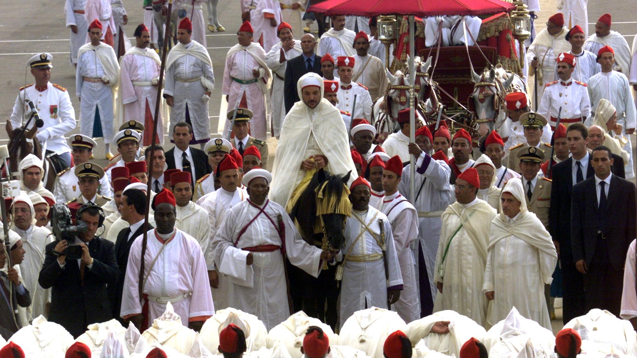 L'image montre un rassemblement cérémonial avec un grand nombre de personnes en tenue traditionnelle. Au centre, une figure importante est portée sur une plate-forme décorée, probablement un leader ou une personnalité éminente. Les participants portent des habits blancs et rouges, indiquant un événement culturel ou religieux. Des officiers en uniformes militaires se trouvent également parmi la foule, et certains photographient la scène. L'ambiance semble festive et respectueuse, typique d'une célébration ou d'une cérémonie officielle.