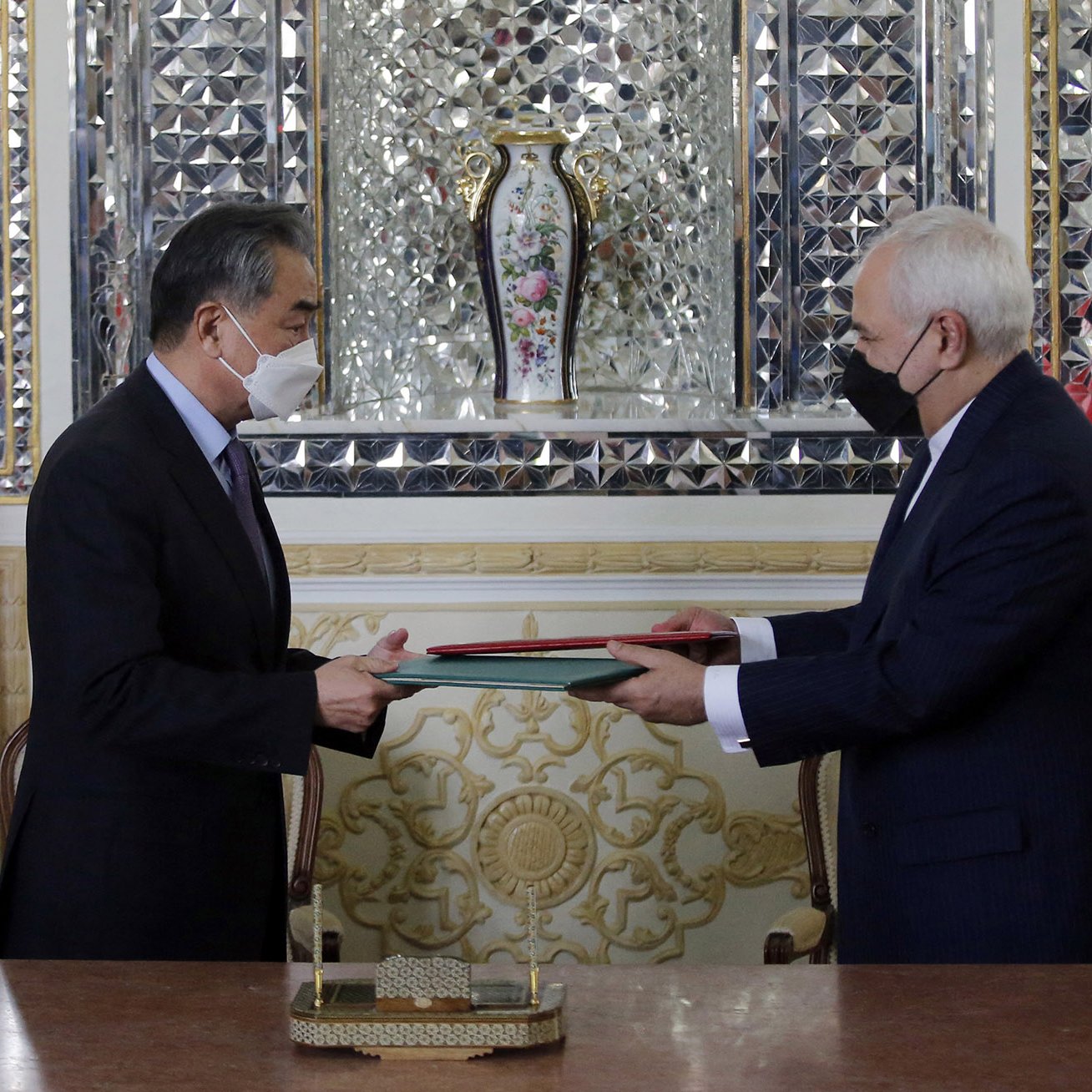 L'image montre deux hommes, apparemment des représentants de la Chine et de l'Iran, se tenant en face l'un de l'autre. Ils échangent des documents au cours d'une cérémonie formelle. On peut voir les drapeaux de la Chine et de l'Iran en arrière-plan. L'environnement est richement décoré, avec des motifs élaborés et un vase floral visible derrière eux. Les deux hommes portent des masques, ce qui pourrait suggérer que la photo a été prise pendant la pandémie.