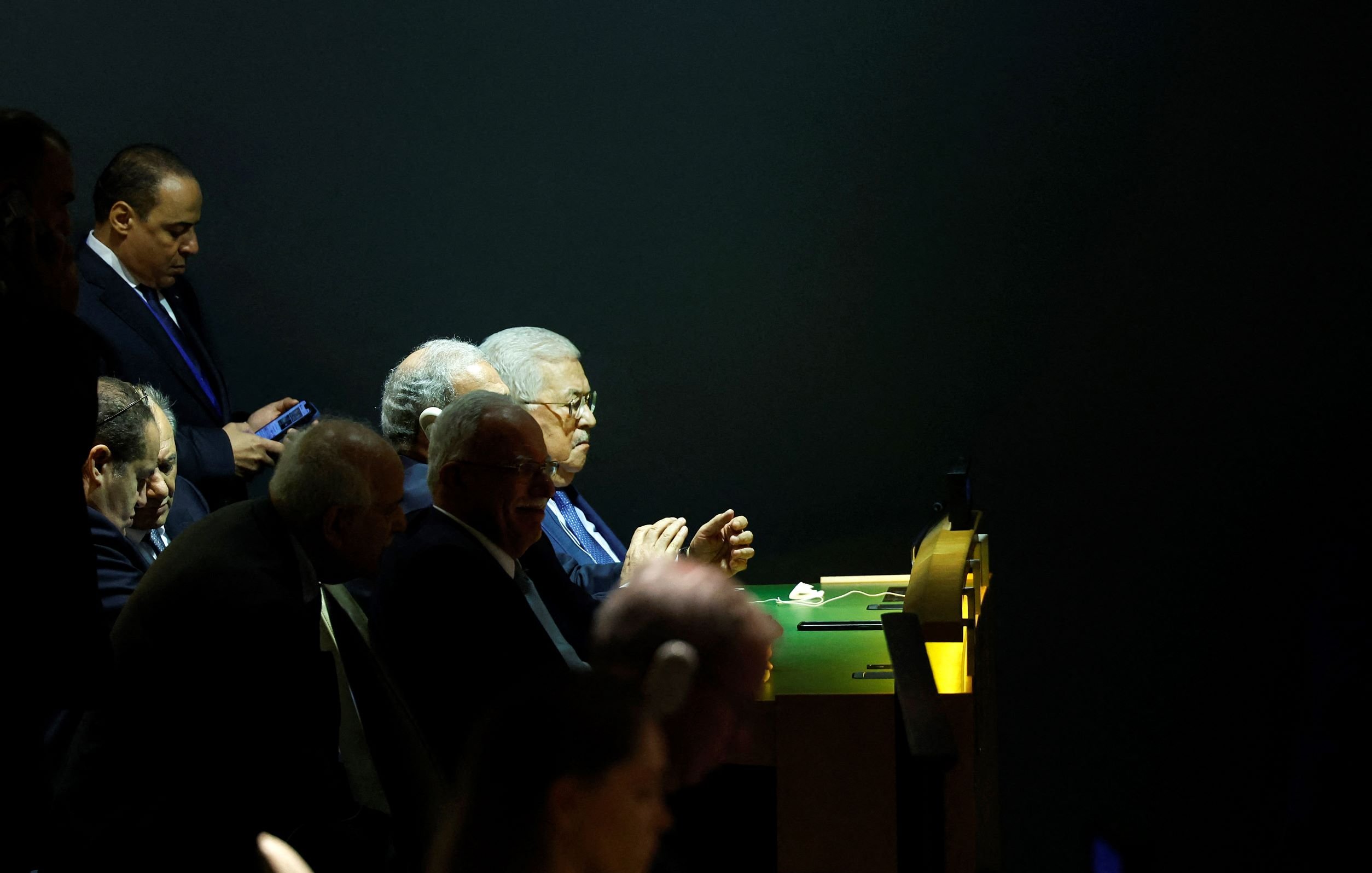 در این تصویر، گروهی از مردان در یک محیط تاریک نشسته‌اند. برخی از آنها در حال صحبت هستند و تمرکز دارند. در میان آنها، فردی با عینک و موهای خاکستری توجه بیشتری را جلب می‌کند. یکی از افراد نیز در حال استفاده از گوشی موبایل خود است. فضای اطراف شامل نورهای ملایمی است که بر روی صحنه یا میز قرار دارد. این تصویر فضایی رسمی و جدی را القا می‌کند.