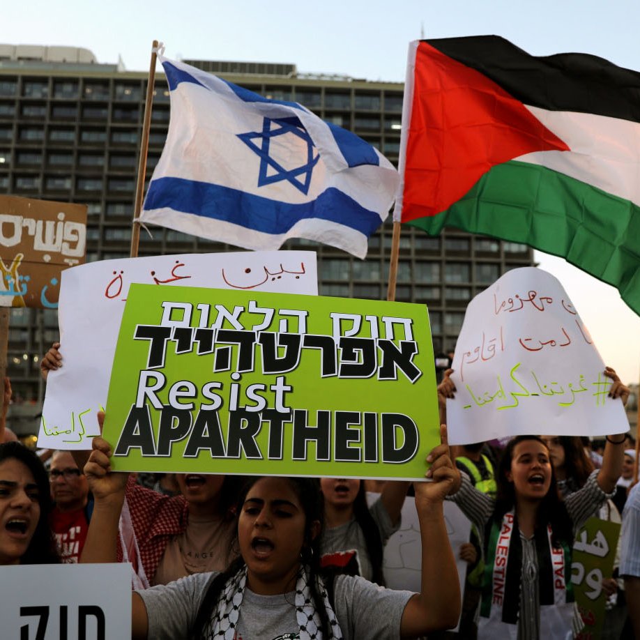 L'image montre une manifestation où des gens tiennent des pancartes. Certains brandissent le drapeau israélien, tandis que d'autres agitent le drapeau palestinien. Les manifestants semblent exprimer des messages de résistance, avec des signes qui dénoncent l'apartheid. L'atmosphère est chargée d'émotion, reflet des tensions et des revendications politiques en cours.