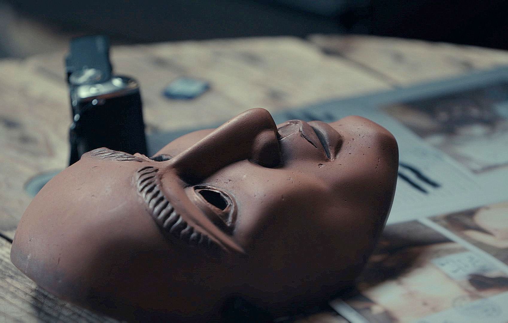L'image montre un masque en terre cuite posé sur une surface en bois. Le masque a des traits détaillés, avec une expression neutre et des sourcils sculptés. À côté du masque, on voit une montre ou un objet similaire, ainsi qu'un magazine ouvert avec des illustrations. L'éclairage est doux, créant une ambiance contemplative autour de ces objets.