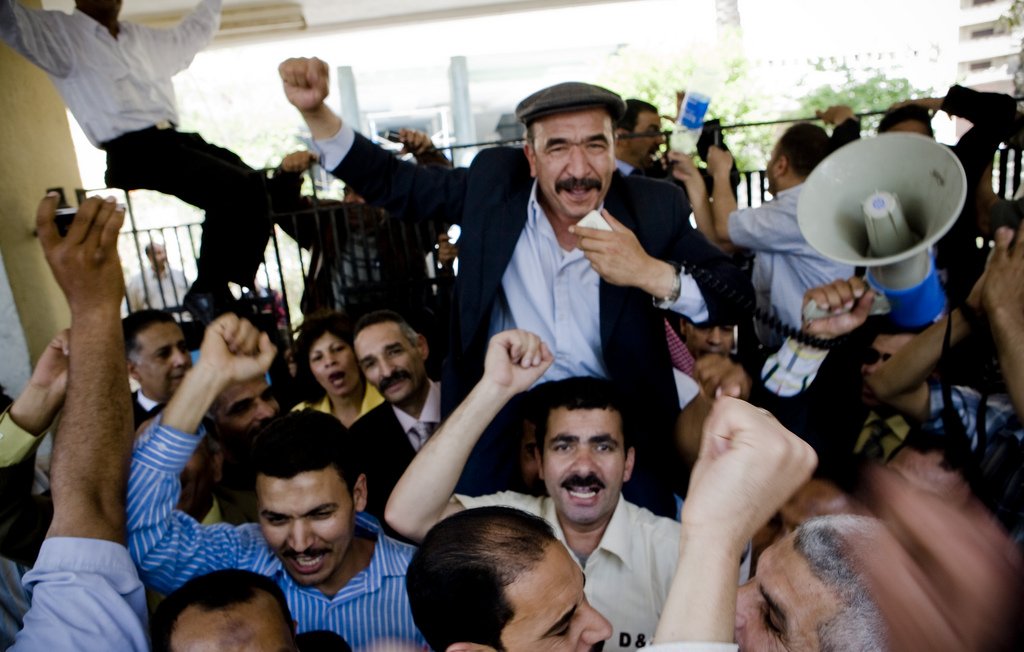 L'image montre une grande foule de personnes rassemblées, manifestant avec enthousiasme. Au centre, un homme souriant est porté sur les épaules d'un autre, levant le poing en signe de victoire. Les autres participants expriment également leur excitation, certains levant les bras et criant. La scène semble vibrante et dynamique, indiquant un moment de célébration ou de rassemblement collectif. Des panneaux et un mégaphone sont également visibles, suggérant une forme de discours ou de protestation.
