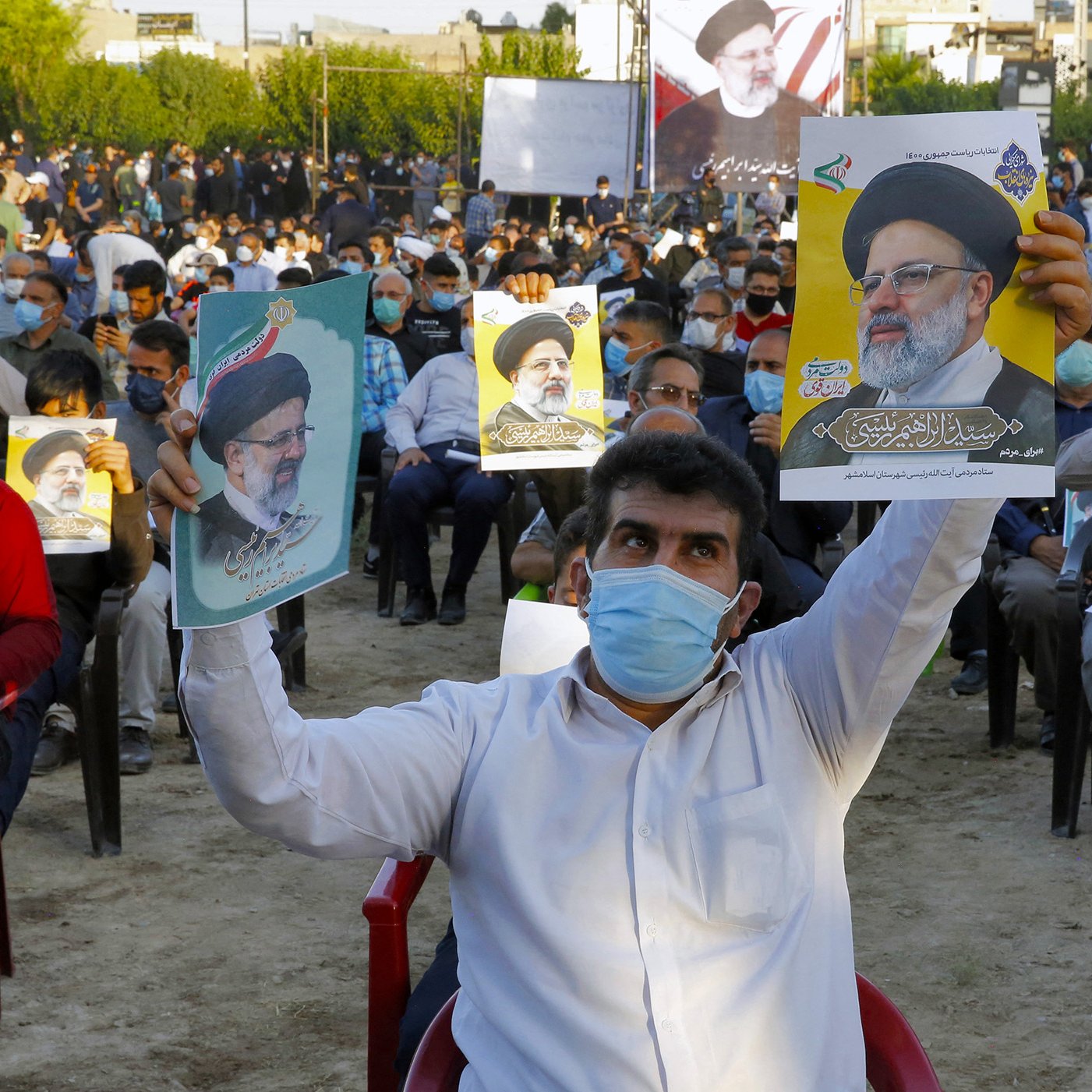 در تصویر، جمعیتی از افراد در یک فضای باز دیده می‌شود که روی صندلی نشسته‌اند و تعدادی از آن‌ها در حال نگه‌داشتن پوسترهایی از یک شخصیت سیاسی هستند. مردم ماسک به‌صورت دارند و در پس‌زمینه، تعدادی پرچم و تابلوهای نمایشی نیز وجود دارد. این جمعیت به نظر می‌رسد که در یک رویداد یا حمایت سیاسی شرکت کرده‌اند.