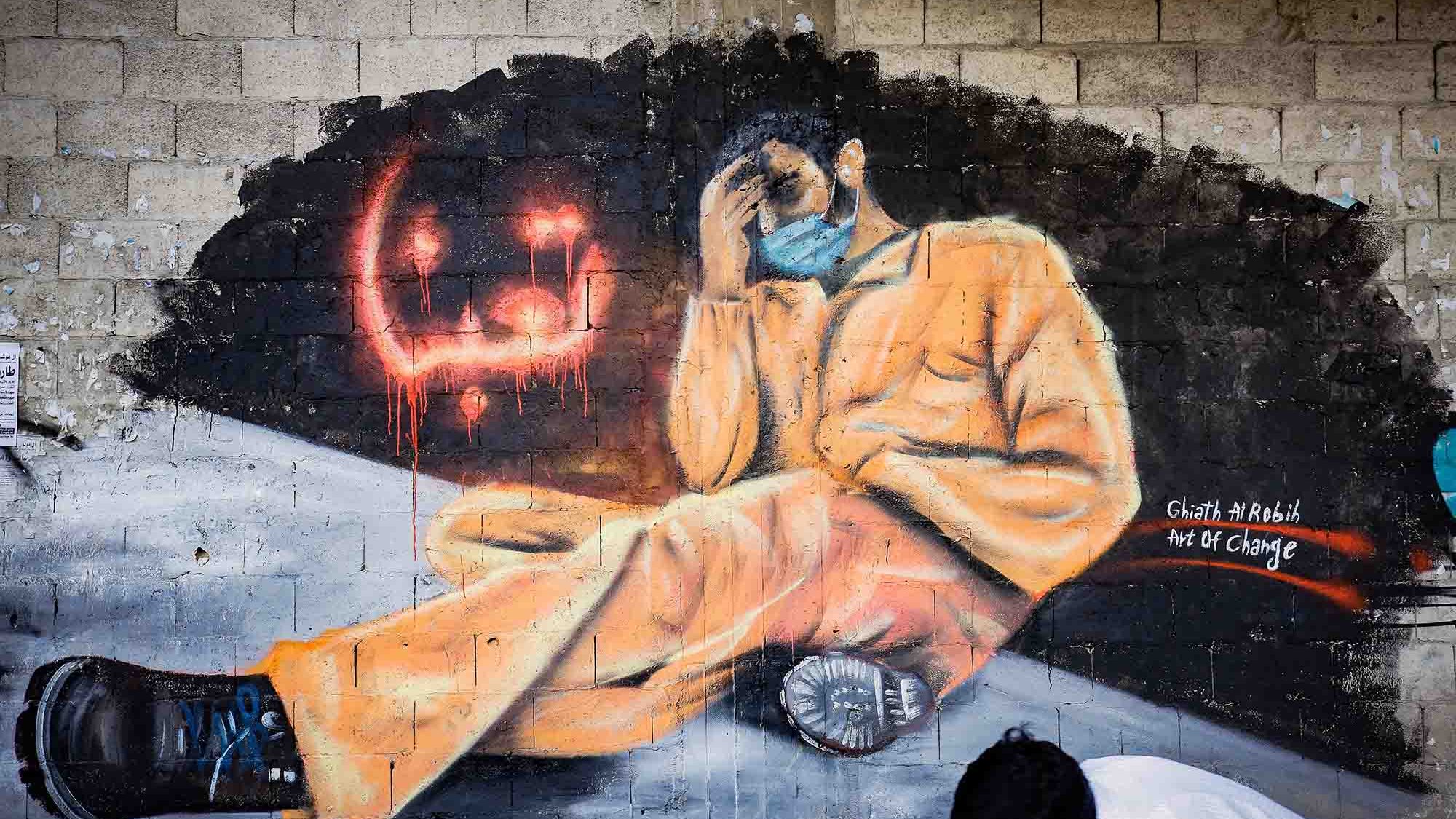 L'image représente une fresque murale colorée montrant un homme assis, habillé d'une combinaison orange. Il semble pensif, la main sur le visage, dans une posture qui évoque la détresse ou la réflexion. Au fond, on aperçoit des graffiti en rouge qui ajoutent une ambiance dramatique à l'œuvre. Un enfant, portant un masque, est également visible en bas de l'image, probablement en train d'observer ou d'interagir avec la fresque. L'ensemble transmet un message puissant lié à des thèmes de changement ou de lutte.