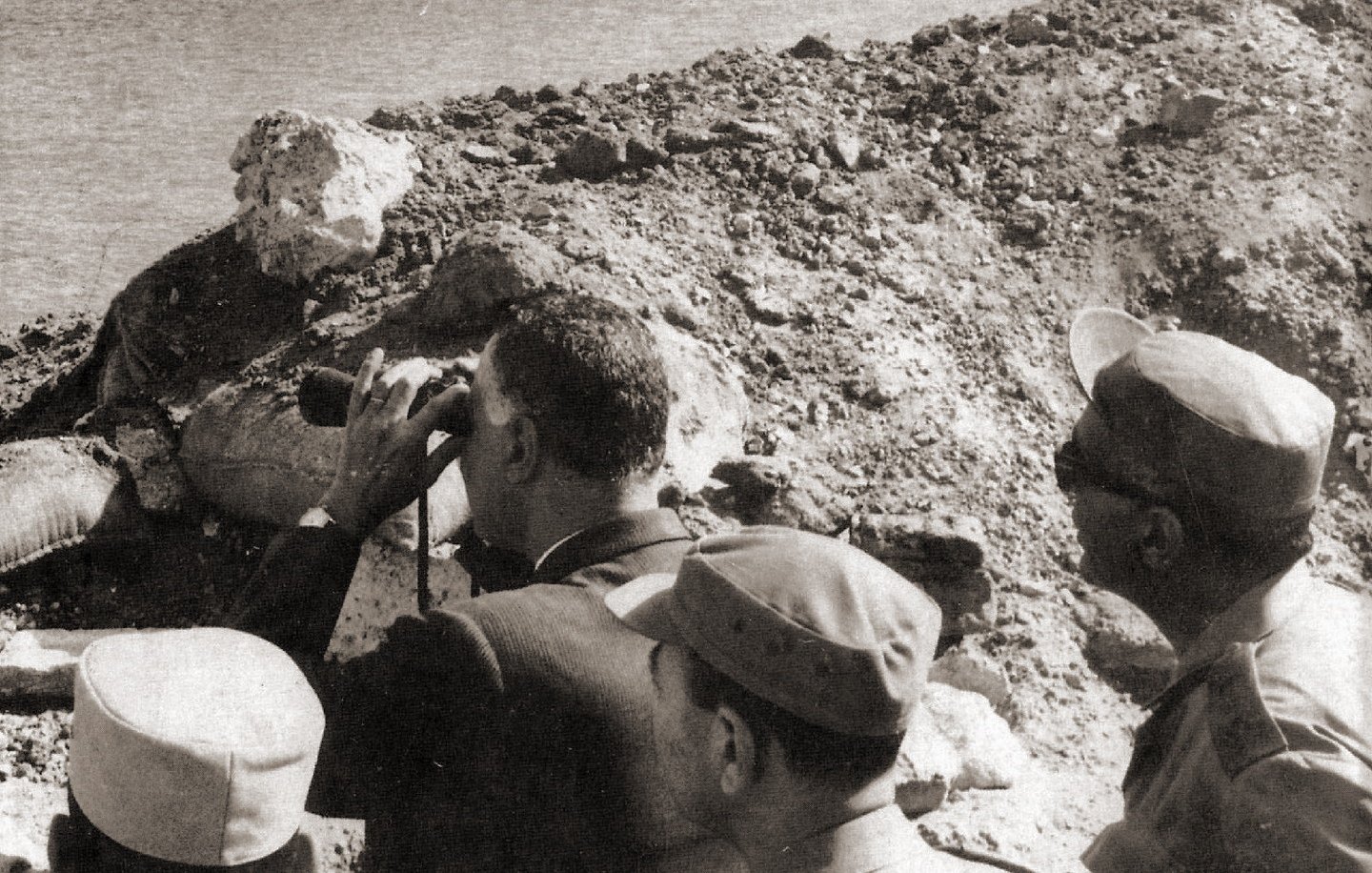 L'image montre un groupe d'hommes observant un paysage, probablement au bord d'une rivière ou d'un lac. L'un des hommes, qui semble en costume, utilise des jumelles pour scruter l'horizon. Les autres sont en tenue militaire et semblent attentifs à ce qui se passe devant eux. L'environnement est aride, avec des collines de terre et peu de végétation visible en arrière-plan. Cette scène évoque une situation de surveillance ou d'évaluation d'un site stratégique.