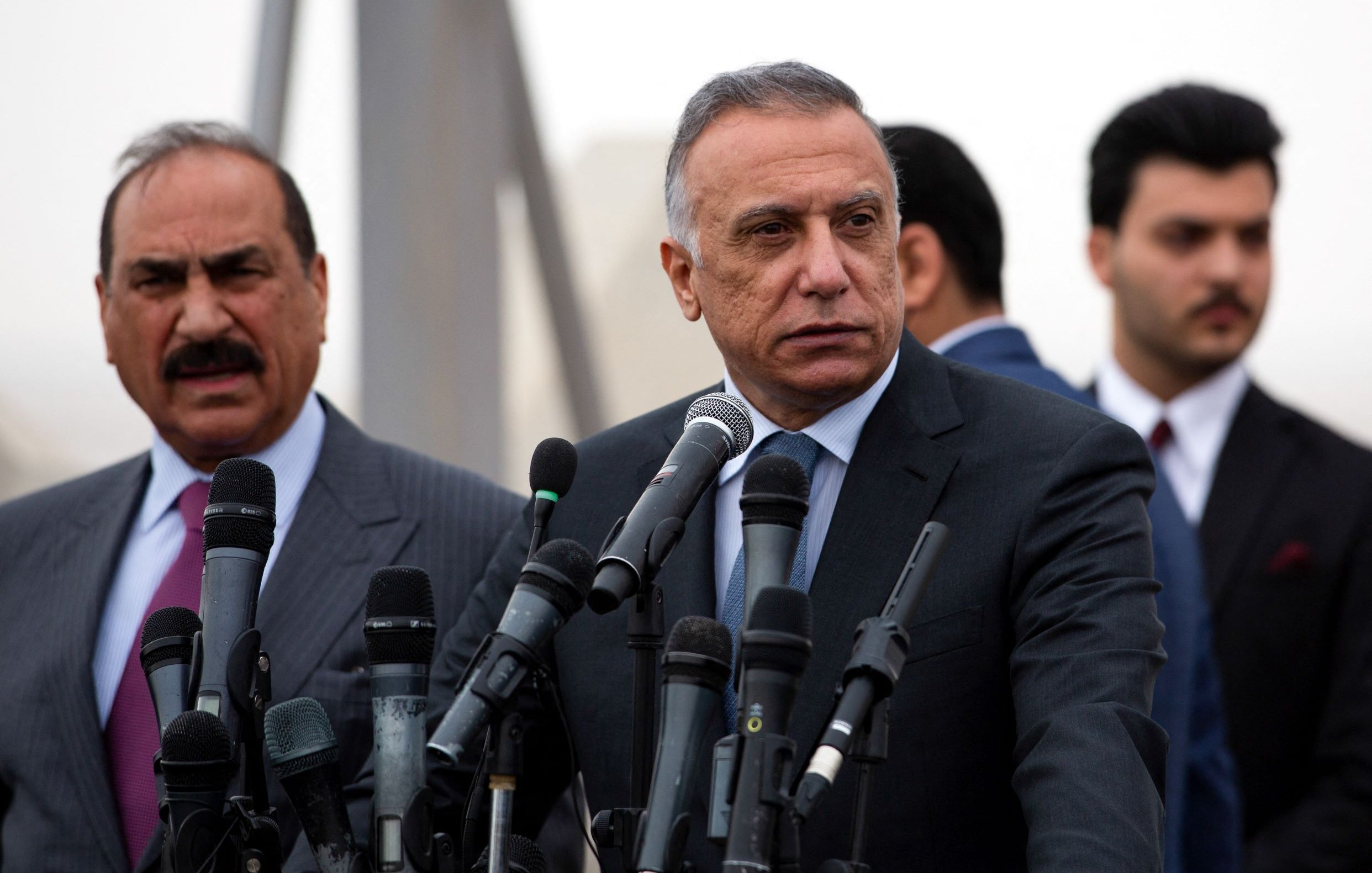 L'image montre un groupe d'hommes lors d'une conférence de presse. Au premier plan, deux hommes sont debout devant plusieurs microphones. L'un d'eux, qui se tient à gauche, porte un costume rayé et semble s'adresser à l'audience. L'autre homme, au centre, est également en costume et adopte une pose sérieuse, regardant au loin. En arrière-plan, d'autres hommes en costumes sont visibles, ajoutant à l'ambiance formelle de l'événement. L'environnement semble légèrement brumeux.