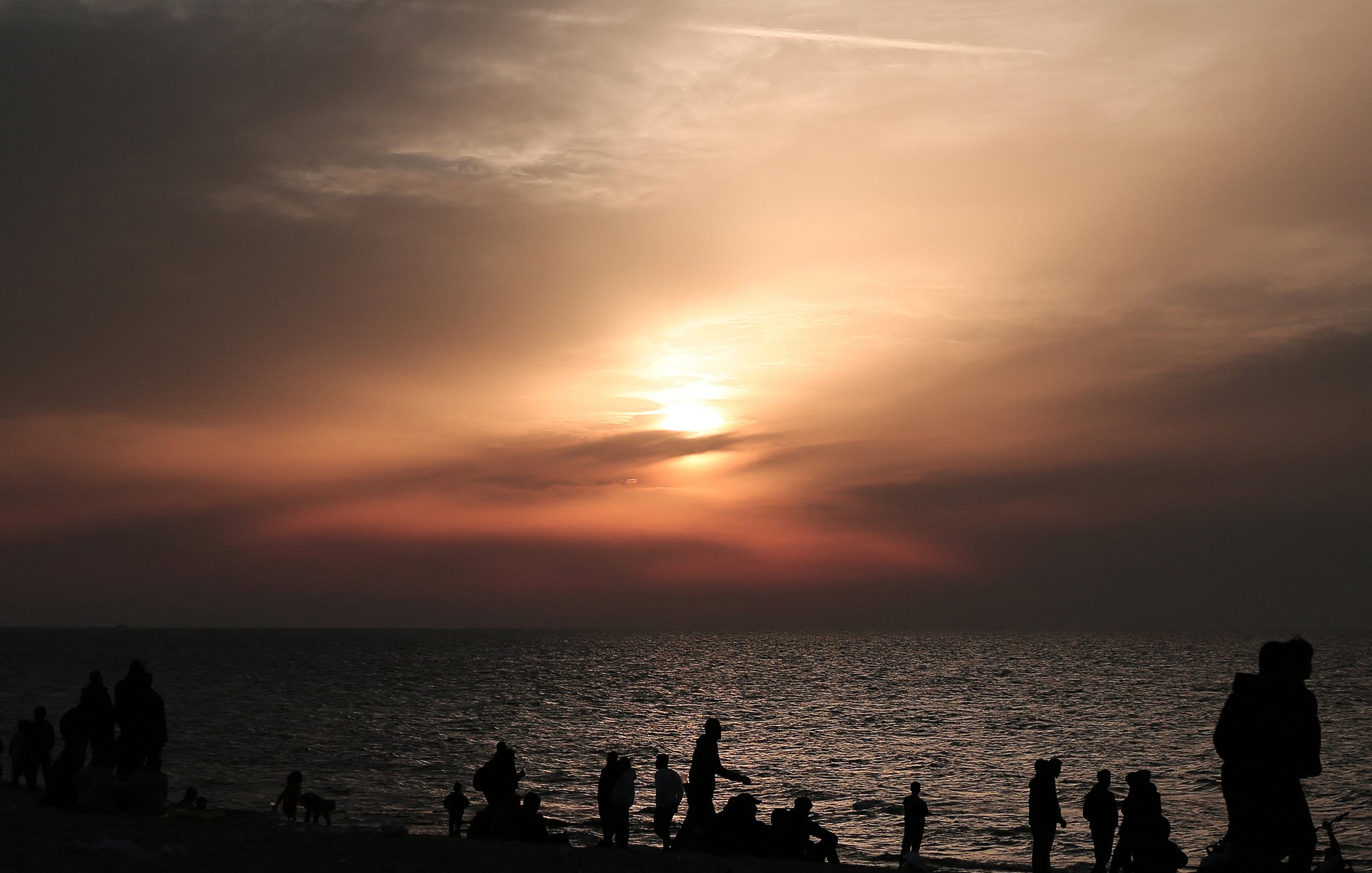 L'immagine mostra una scena al tramonto sulla spiaggia. Il cielo è dipinto di tonalità calde, con sfumature di arancione e rosa, mentre il sole si avvicina all'orizzonte. In primo piano, si vedono diverse silhouette di persone che camminano lungo la riva, alcune sedute o accoccolate sulla sabbia. La superficie dell'acqua riflette i colori del cielo, creando un'atmosfera calma e suggestiva.