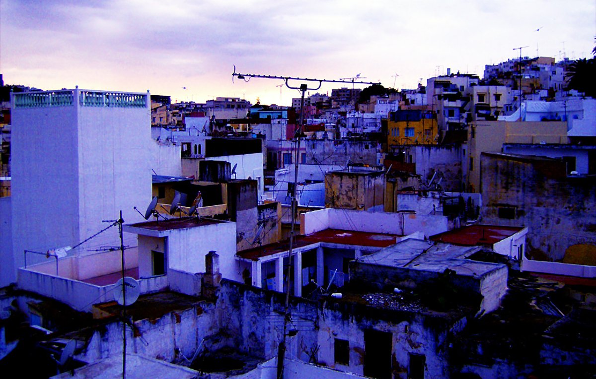 L'image montre une vue urbaine d'un paysage de ville, probablement à la tombée de la nuit. On peut voir plusieurs bâtiments aux structures variées, avec des toits plats et un mélange de couleurs qui évoquent une atmosphère un peu mélancolique. Certains bâtiments semblent légèrement en ruine, avec des murs décrépis et des débris visibles. Le ciel est nuageux, ajoutant une ambiance dramatique à la scène. Des antennes de télévision sont visibles sur les toits, typiques des environnements urbains. L'ensemble donne une impression de vie quotidienne dans une ville qui a du caractère.
