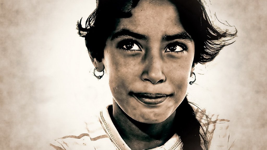 L'image montre un portrait d'une jeune fille. Elle a un regard pensif avec une légère expression sur son visage. Ses cheveux sont sombres et légèrement ébouriffés, et elle porte des boucles d'oreilles. Le fond est flou, mettant en valeur ses traits. Les contrastes de l'image donnent une atmosphère intense et émotive.