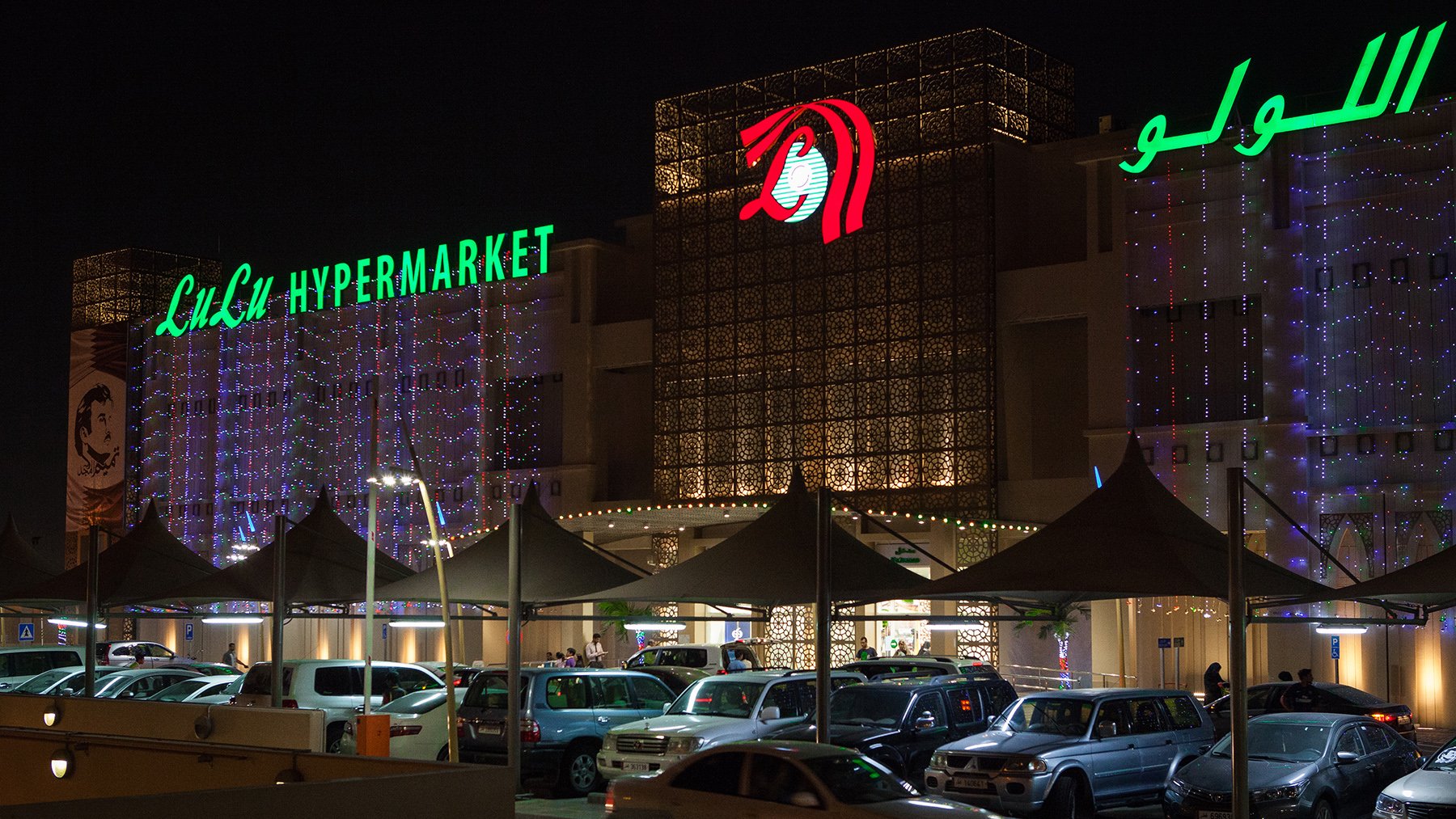 تظهر الصورة واجهة مركز تجاري مضاء في الليل، مع لافتات بارزة تحمل اسم "لولو هايبرماركت". التصميم الخارجي يتضمن أضواء زاهية وزخارف جميلة، مما يضفي طابعاً مبهجاً على المكان. كما يوجد أمام المركز مواقف للسيارات، تُظهر وجود عدد من السيارات المتعددة الألوان. الجو العام يوحي بالحيوية والنشاط، مع وجود بعض الأشخاص حول المدخل.