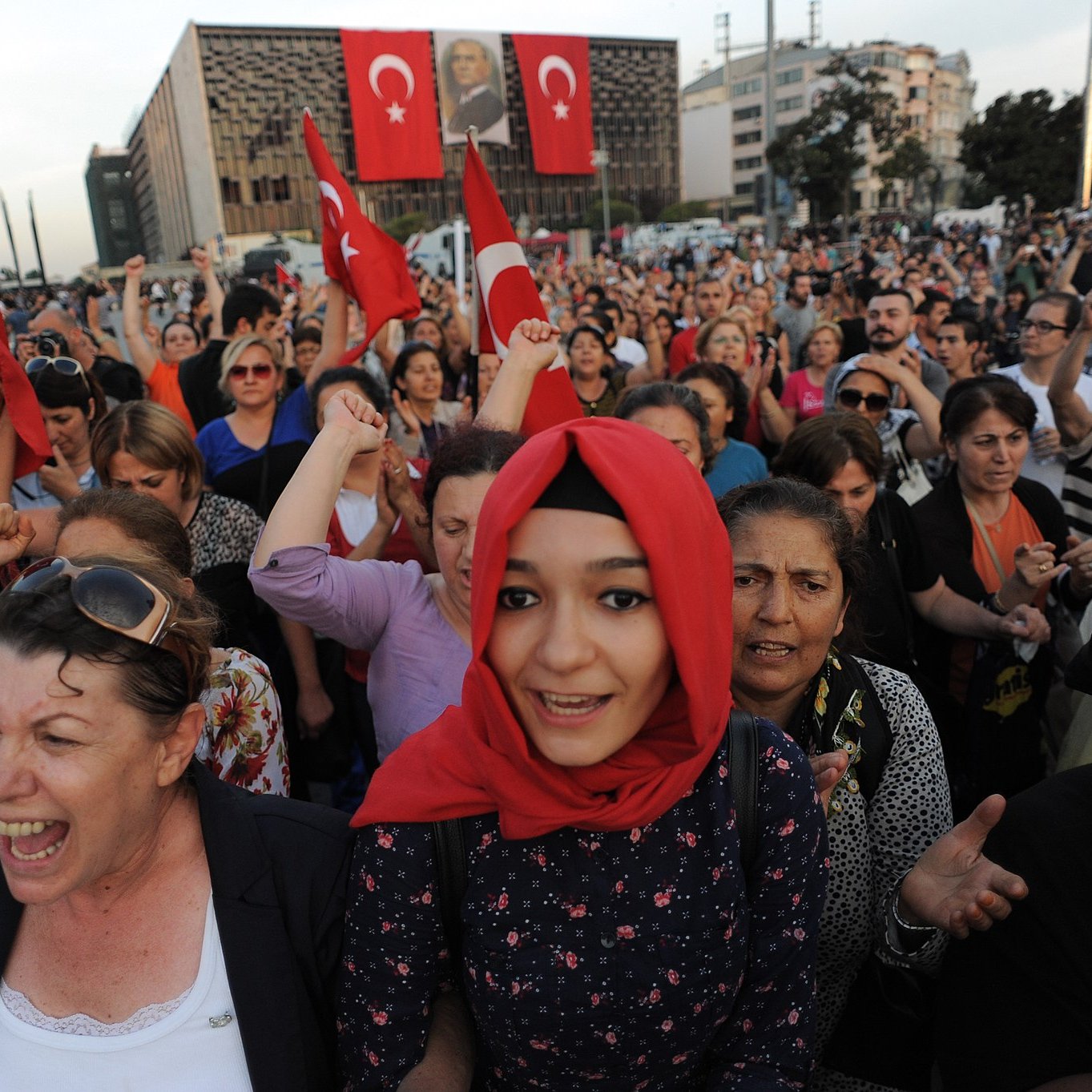 The image depicts a large crowd gathered, likely in an outdoor setting, with many individuals holding Turkish flags. People appear to be passionately engaged, with expressions of enthusiasm and solidarity. Some individuals are wearing traditional attire, while others wear modern clothing. The background features a building adorned with large banners, adding to the sense of national pride and collective emotion in the scene. The atmosphere seems charged with energy, suggesting a demonstration or celebration of national significance.