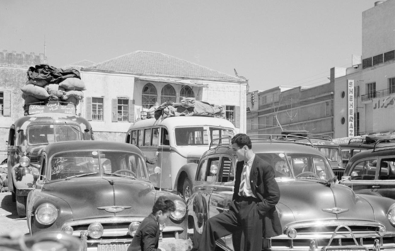 L'image montre une scène de rue animée, probablement dans un contexte urbain d'une époque passée. Au premier plan, un homme en costume se tient debout, l'air décontracté, avec un pied posé sur une petite caisse. Devant lui, un autre homme est accroupi, occupé à préparer quelque chose dans un petit pot ou une casserole. En arrière-plan, on peut voir de nombreuses voitures anciennes, avec des bagages sur leur toit, suggérant peut-être un moment de transition ou de voyage. Le cadre est lumineux et semble capter une ambiance d'activité dynamique, typique des villes dans les décennies passées.