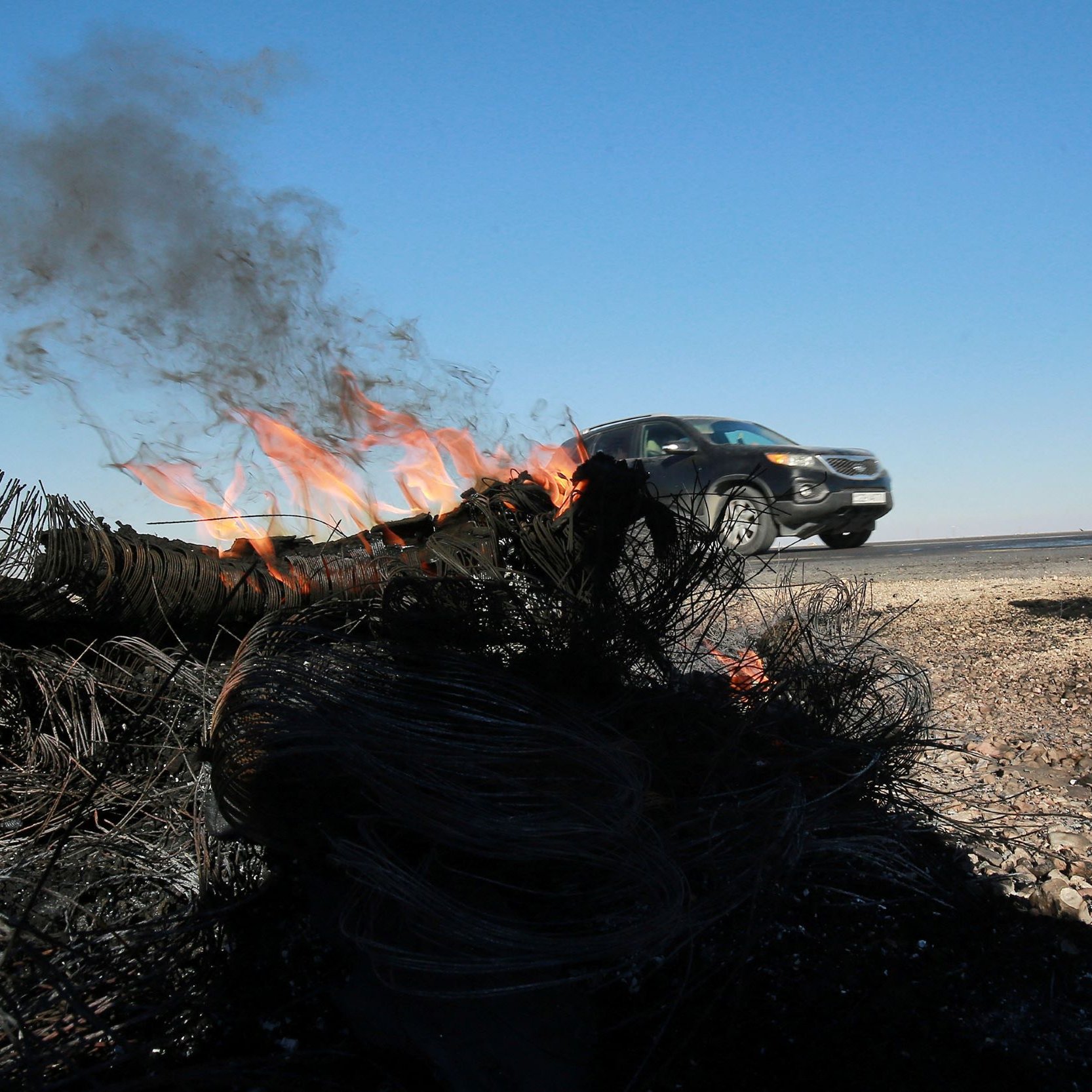 L'image montre un véhicule qui roule rapidement le long d'une route, tandis qu'un feu brûle sur le bord de la route. De la fumée s'élève du feu, qui semble être alimenté par des matériaux tels que des câbles ou des filets. Le ciel est dégagé avec une couleur bleue, et le paysage environnant est aride. On peut voir des débris brûlés au premier plan, ce qui suggère une scène de destruction ou d'accident.