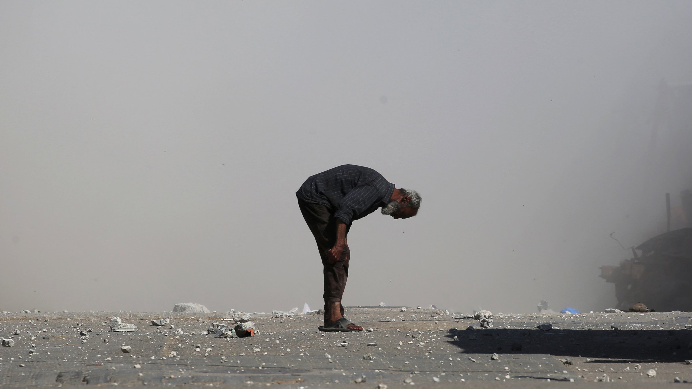 A man bends over on a dusty street, surrounded by debris and smoke.