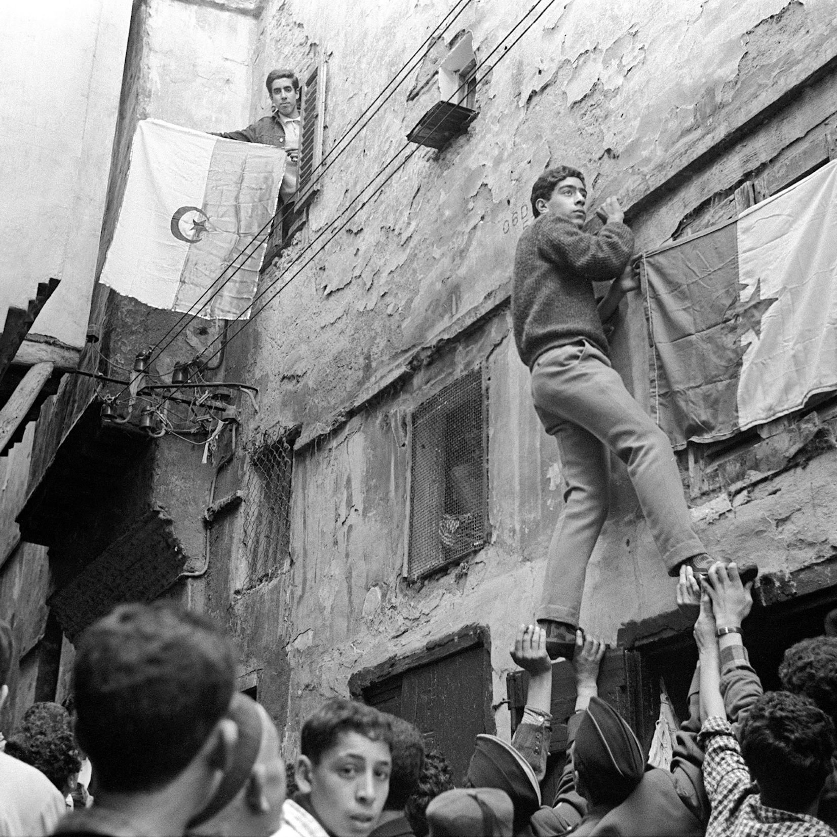 L'image montre une scène dynamique dans une ruelle animée. Des personnes rassemblées en bas soutiennent quelqu'un qui grimpe le long d'un mur. Ce dernier semble s'accrocher à un drapeau, tandis qu'un autre drapeau est visible au-dessus, accroché à une fenêtre. Les murs de l'immeuble sont en mauvais état, ajoutant une atmosphère de tension historique à la scène. Les visages des personnes expriment un mélange d'espoir et de détermination. Cette image évoque un moment significatif lié à des luttes ou des revendications.
