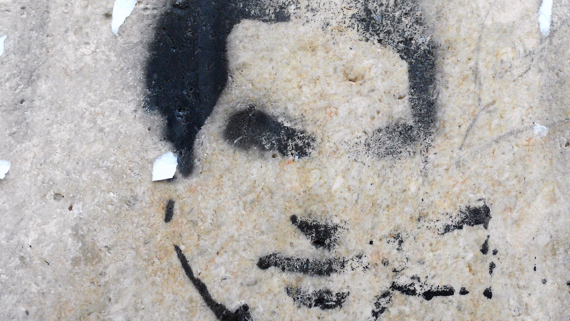 L'image montre un pochoir sur un mur, représentant un visage stylisé. Les contours sont en noir, avec un fond de pierre claire. Le visage semble être celui d'une personne jeune, avec une expression neutre. Quelques résidus de papier sont éparpillés autour, ajoutant une texture supplémentaire au mur. L'ensemble dégage une atmosphère urbaine et artistique.