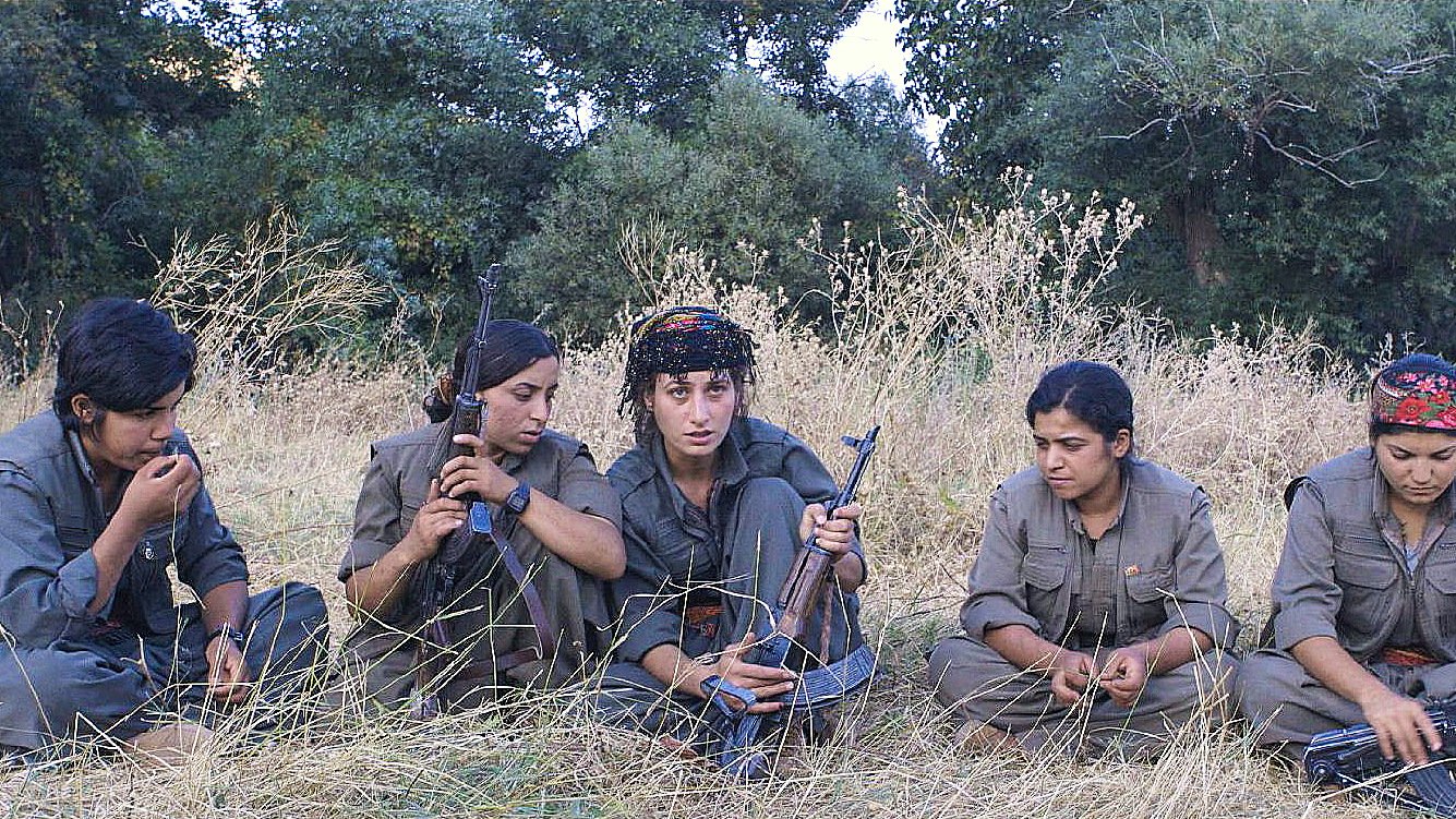 L'image montre un groupe de cinq femmes assises dans une prairie, entourées de végétation. Elles portent des vêtements militaires et semblent concentrées sur une activité commune. Certaines d'entre elles tiennent des armes à feu, tandis que d'autres ont les mains posées sur leurs genoux. Leurs expressions faciales et leur posture suggèrent une ambiance sérieuse et déterminée.