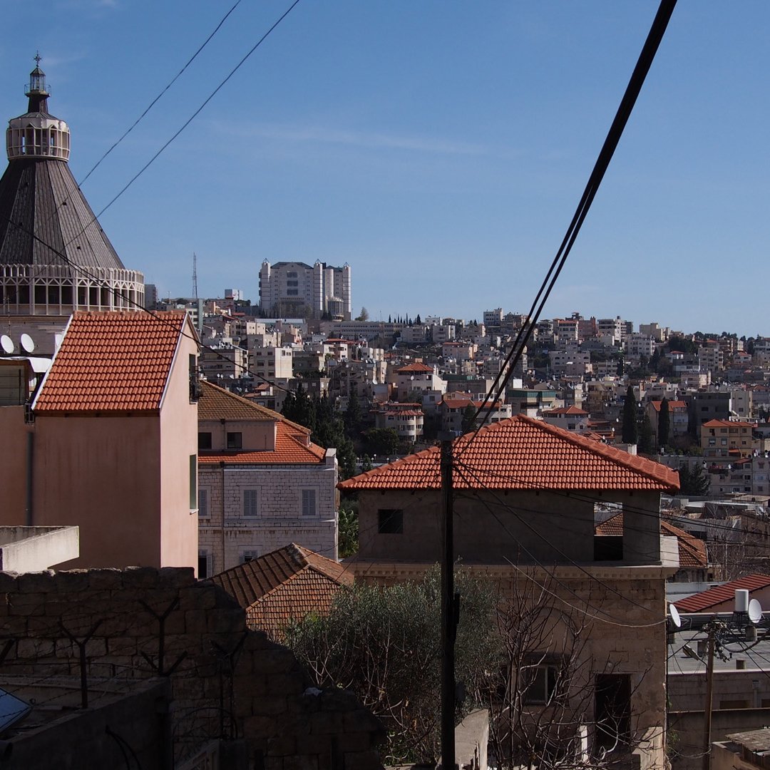 L'immagine mostra un paesaggio urbano con diverse abitazioni disposte su una collina. In primo piano, ci sono edifici con tetti a spiovente di colore rosso e strutture in muratura. Sullo sfondo si riconosce un grande edificio con una copertura a cupola, che si erge tra le case. Il cielo è sereno e limpido, contribuendo a dare un'impressione di vivacità al panorama cittadino. Inoltre, alcuni fili elettrici attraversano l'immagine, evidenziando l'aspetto urbano della scena.