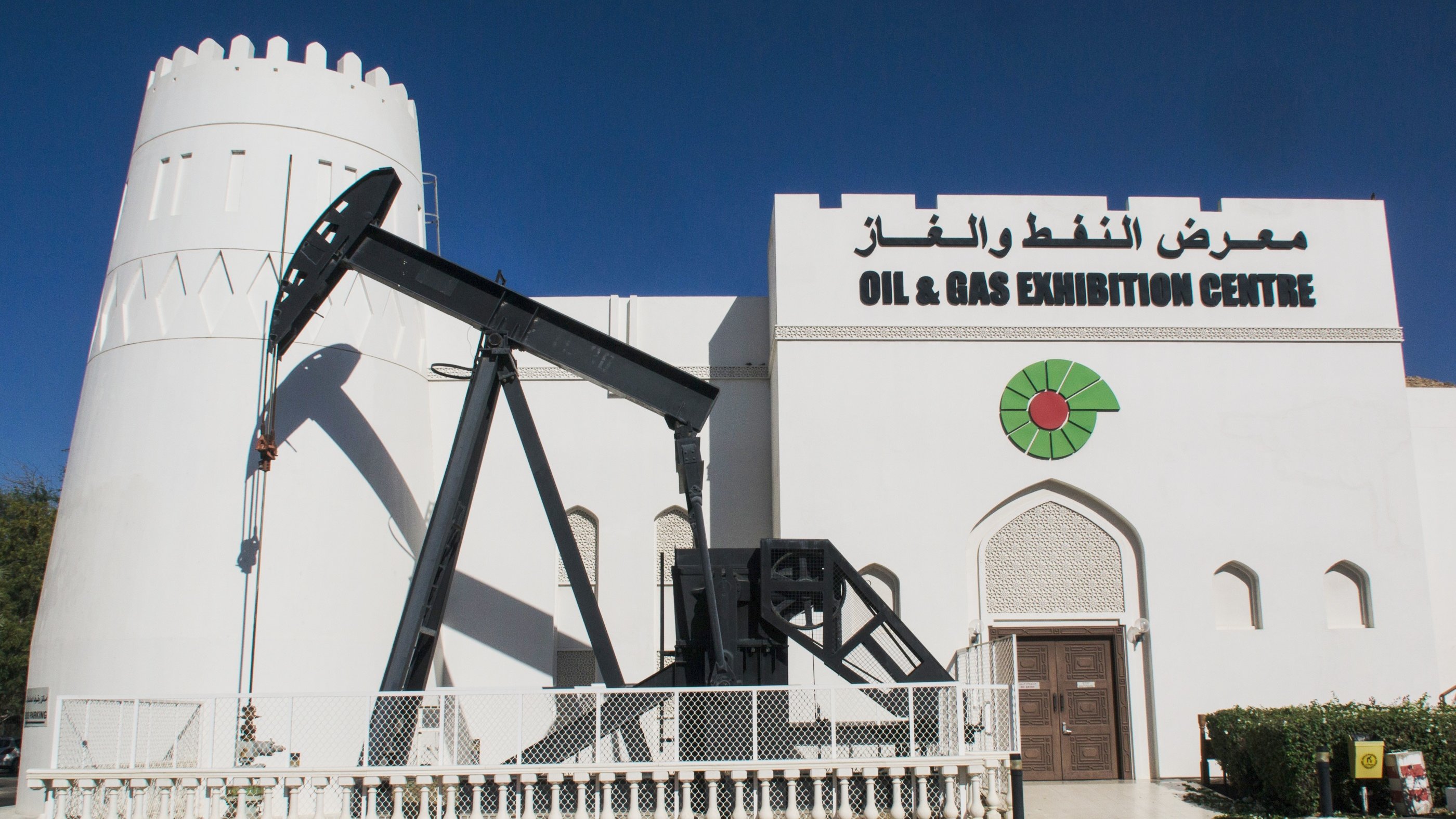 L'image montre un bâtiment blanc qui semble être un centre d'exposition dédié au pétrole et au gaz. À gauche, il y a une structure en forme de pompe à pétrole, typique des installations de forage. Le bâtiment présente un style architectural qui rappelle une forteresse, avec une tour à moitié ronde sur le côté. Au-dessus de l'entrée, on peut voir le nom "OIL & GAS EXHIBITION CENTRE" ainsi que des inscriptions en arabe. La façade est décorée de motifs graphiques, ajoutant une touche esthétique au bâtiment. Le ciel est clair et bleu en arrière-plan.