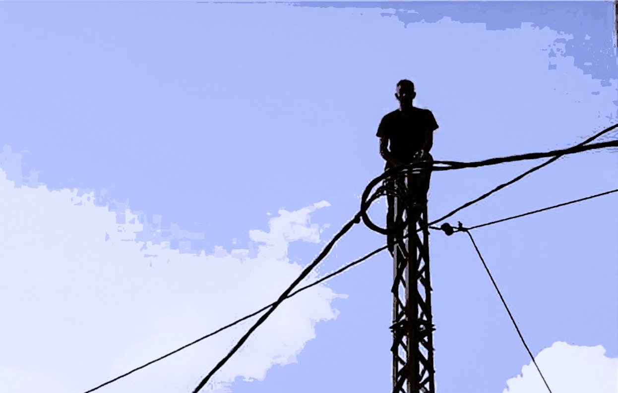 Dans cette image, on peut voir un homme debout sur un pylône, entouré de câbles. Le ciel en arrière-plan est bleu avec quelques nuages. La silhouette de l'homme est mise en avant par le contraste avec le ciel. L'atmosphère semble à la fois audacieuse et impressionnante, soulignant une certaine prise de risque.