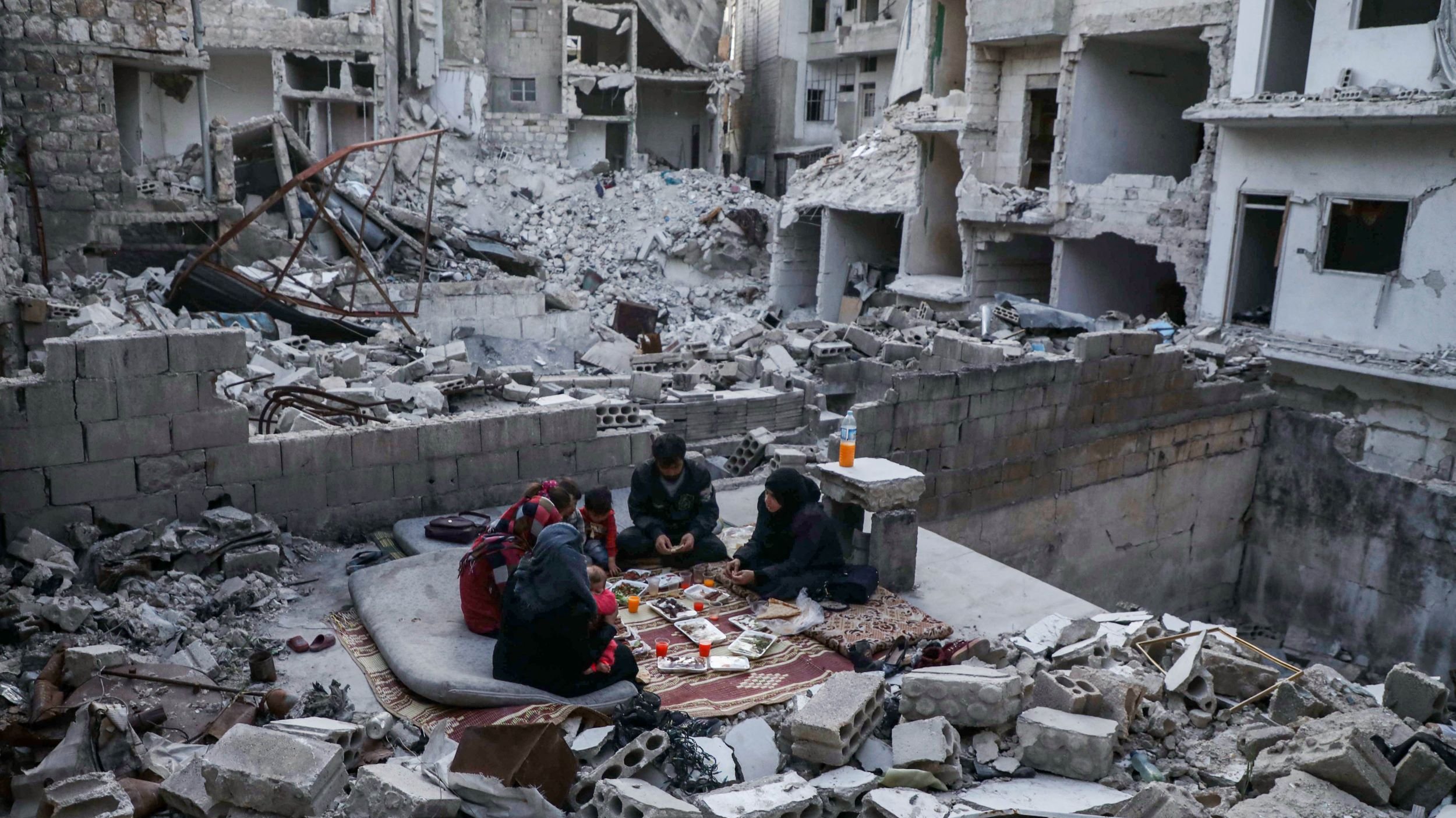 L'immagine mostra un contesto di distruzione, probabilmente in seguito a un conflitto. Al centro, un gruppo di persone è seduto su un tappeto, apparentemente intenti a giocare o a trascorrere del tempo insieme. Attorno a loro ci sono macerie e detriti di edifici crollati, con pareti distrutte e strutture parzialmente in piedi. La scena trasmette un senso di resilienza nel mezzo della devastazione.