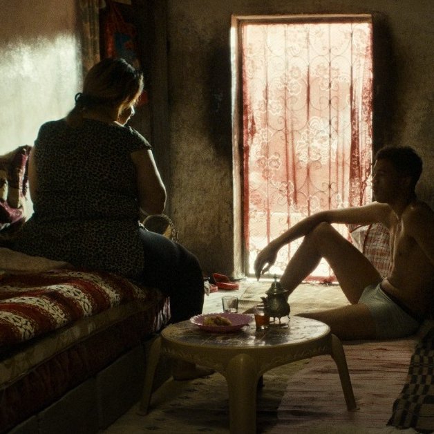 L'image montre une scène intérieure, probablement dans un espace de vie modeste. On y voit deux personnes, assises dans une pièce faiblement éclairée par la lumière qui entre à travers une fenêtre avec des rideaux. Les murs sont de couleur sombre et le mobilier semble simple. L'une des personnes est assise sur un canapé, tandis que l'autre est assise par terre, semblant contempler la pièce ou se relaxer. L'atmosphère dégage une sensation d'intimité et de calme. Des objets sont présents, suggérant une vie quotidienne.