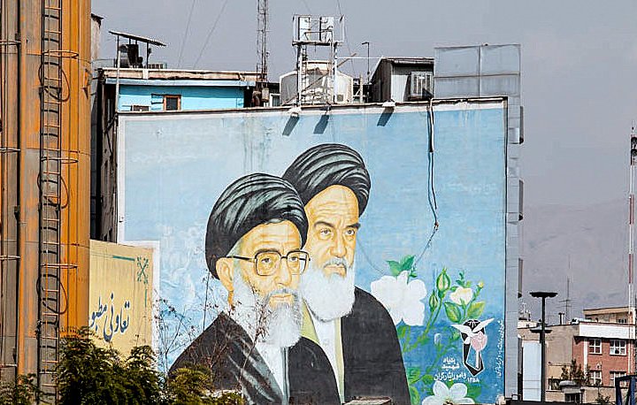 L'image montre une grande fresque murale représentant deux hommes avec des turbans noirs et des barbes blanches. Ils sont peints sur un mur d'un bâtiment, entourés de fleurs et de motifs décoratifs. En arrière-plan, on aperçoit des structures urbaines et un ciel légèrement nuageux. Cette fresque semble symboliser une figure ou un message important dans le contexte culturel ou historique de la région.