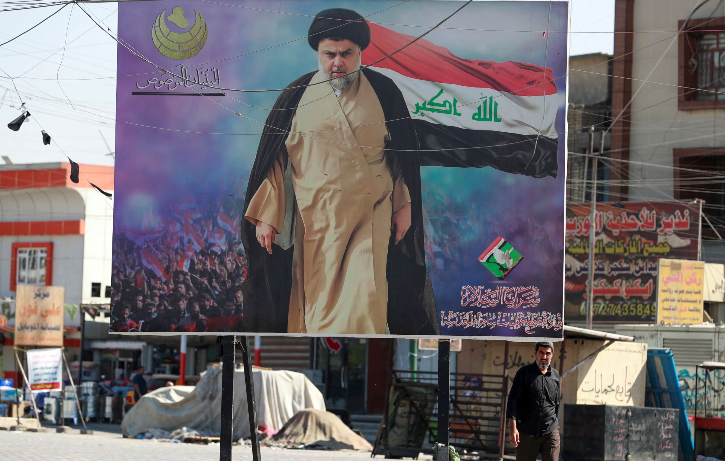 L'image montre une grande affiches publicitaire dans un espace urbain. Au centre, un homme en costume traditionnel, probablement un religieux, est représenté avec un drapeau. Le fond de l'affiche semble comporter une foule, ce qui suggère un événement ou un rassemblement. Une personne marche devant l'affiche, ajoutant une dimension humaine à la scène. Les couleurs dominantes sont le noir, le rouge et le vert, qui sont souvent symboliques dans le contexte politique.