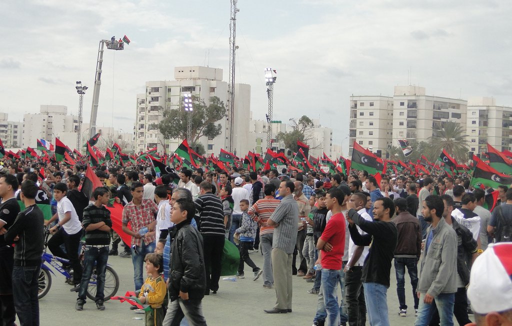 L'image montre une foule rassemblée dans un espace public, probablement lors d'un événement ou d'une célébration. De nombreuses personnes, dont des hommes, des femmes et des enfants, tiennent des drapeaux aux couleurs rouge, noir et vert, qui sont typiques du drapeau libyen. L'ambiance semble festive et dynamique, avec des bâtiments modernes en arrière-plan et des équipements techniques pour la sonorisation ou l'éclairage. On peut également voir des personnes sur des vélos, ajoutant une dimension de vie quotidienne à la scène.