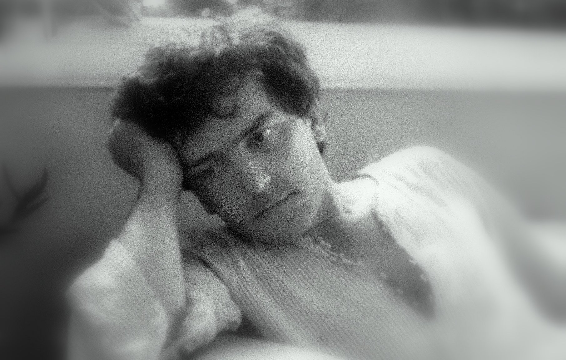 L'image montre un jeune homme pensif, appuyé sur sa main, avec une expression mélancolique. Il semble perdu dans ses pensées, ses cheveux sont légèrement ondulés et il porte une chemise à manches longues. L'arrière-plan est flou, créant une ambiance douce et introspective. La lumière est tamisée, ajoutant à l'atmosphère de réflexion.