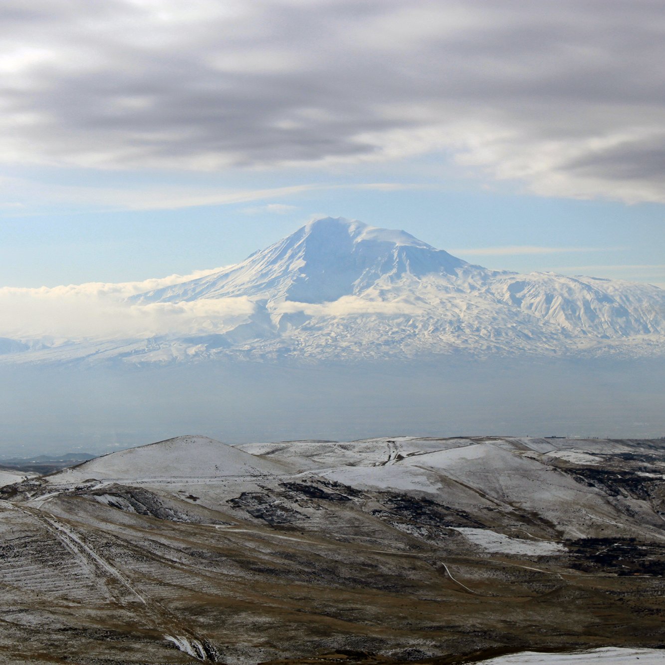 L'image montre un paysage montagneux impressionnant, avec un grand sommet enneigé qui semble être un volcan. Le sommet est entouré de nuages légers, créant une atmosphère mystique. Au premier plan, des collines aux teintes terreuses et parfois enneigées s'étendent, ajoutant à la beauté naturelle de la scène. Le ciel est partiellement couvert, laissant transparaître une lumière douce qui éclaire le paysage.