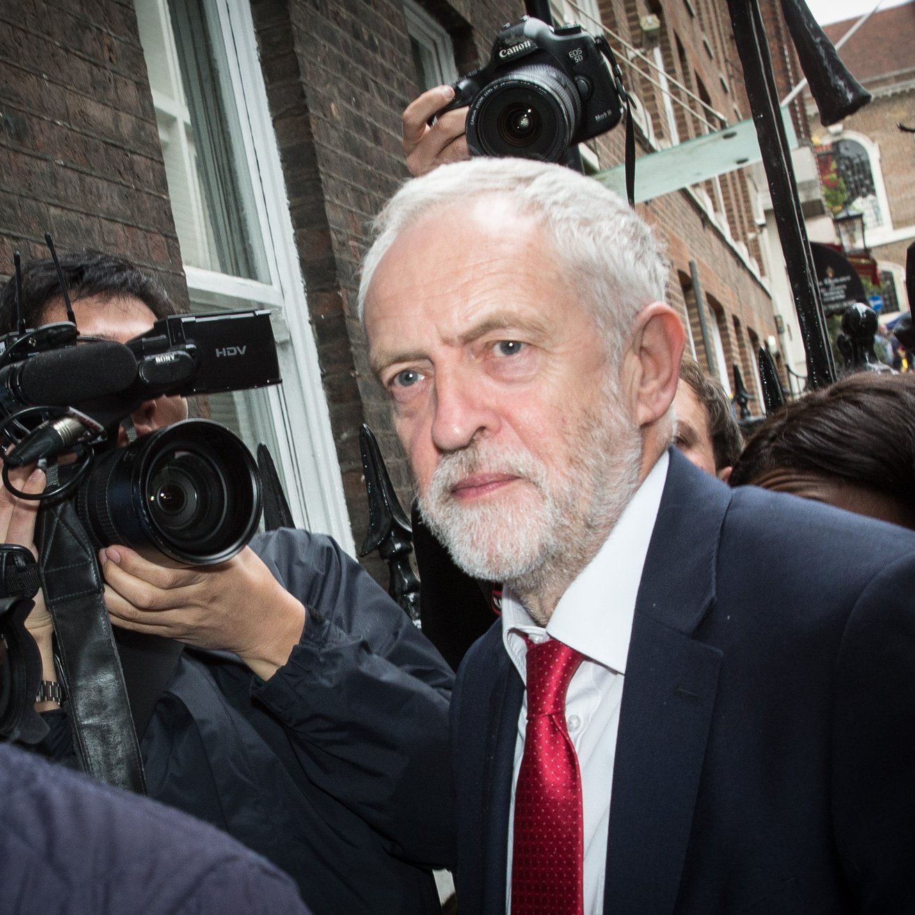 L'image montre un homme avec des cheveux gris et une barbe, vêtu d'un costume sombre et d'une cravate rouge. Il semble se déplacer dans un environnement urbain, entouré de journalistes et de caméras qui l'interrogent ou le photographient. L'atmosphère est animée et il est probable qu'il soit au centre de l'attention médiatique. Le cadre présente des bâtiments en briques, suggérant une ambiance citadine.