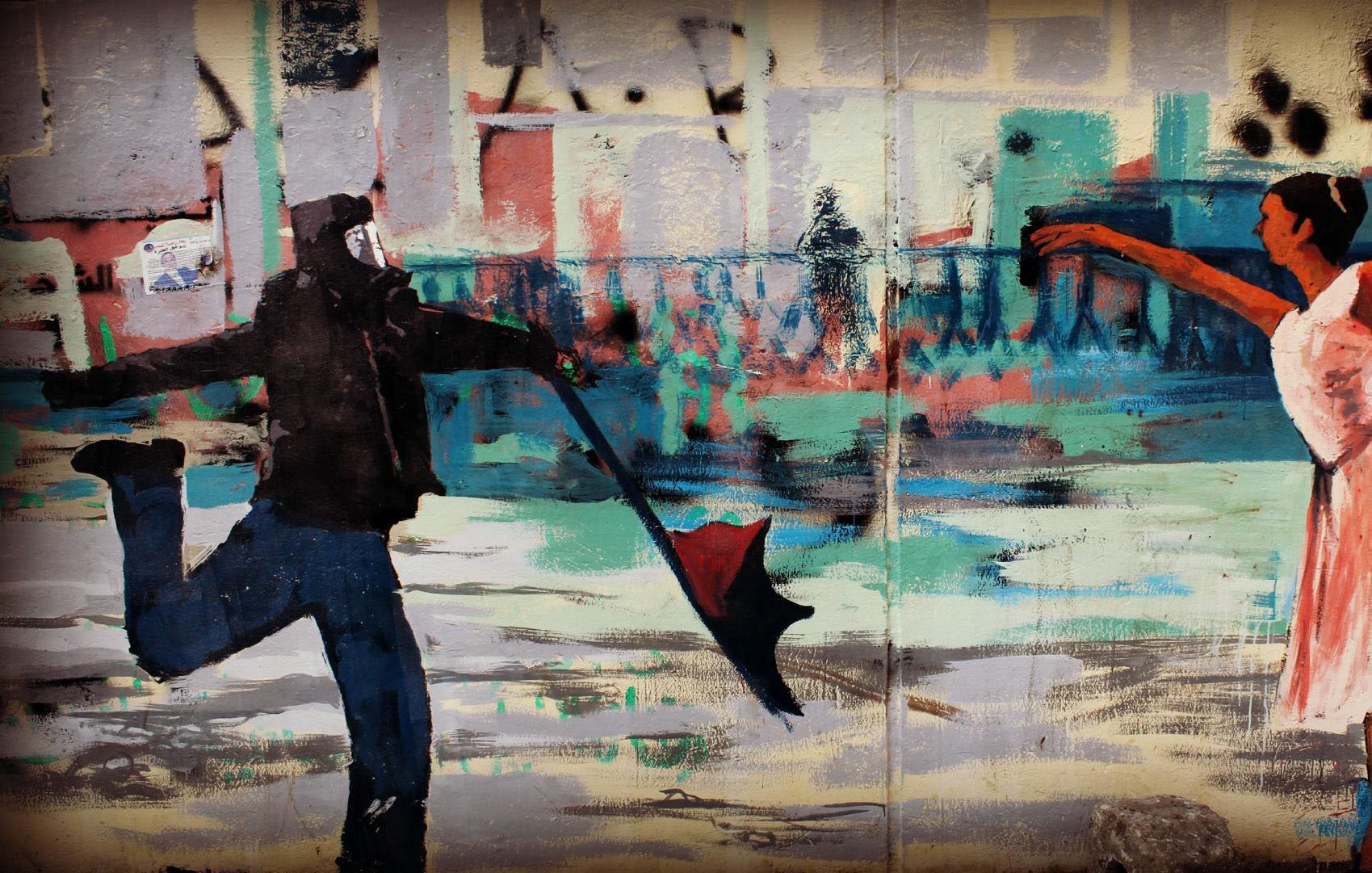 L'image montre une œuvre de street art. À gauche, on voit un homme portant un masque, habillé de noir, qui tient un parapluie ouvert. À droite, une ballerine en robe blanche exécute une danse gracieuse. Le fond de l'œuvre est rempli de couleurs vives et de formes abstraites, créant une atmosphère dynamique. Cette juxtaposition entre les deux personnages évoque un contraste entre l'agitation du monde urbain et la beauté de la danse.