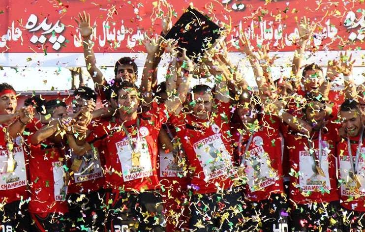 L'image montre une équipe de football célébrant une victoire. Les membres de l'équipe portent des maillots rouges et affichent des sourires de joie. Ils tiennent des trophées et sont entourés de confettis colorés qui tombent sur eux. L'ambiance est festive, et on peut sentir l'excitation de la victoire à travers leurs expressions. Des banderoles et des affiches derrière eux montrent le nom de l'équipe et le mot "champion".