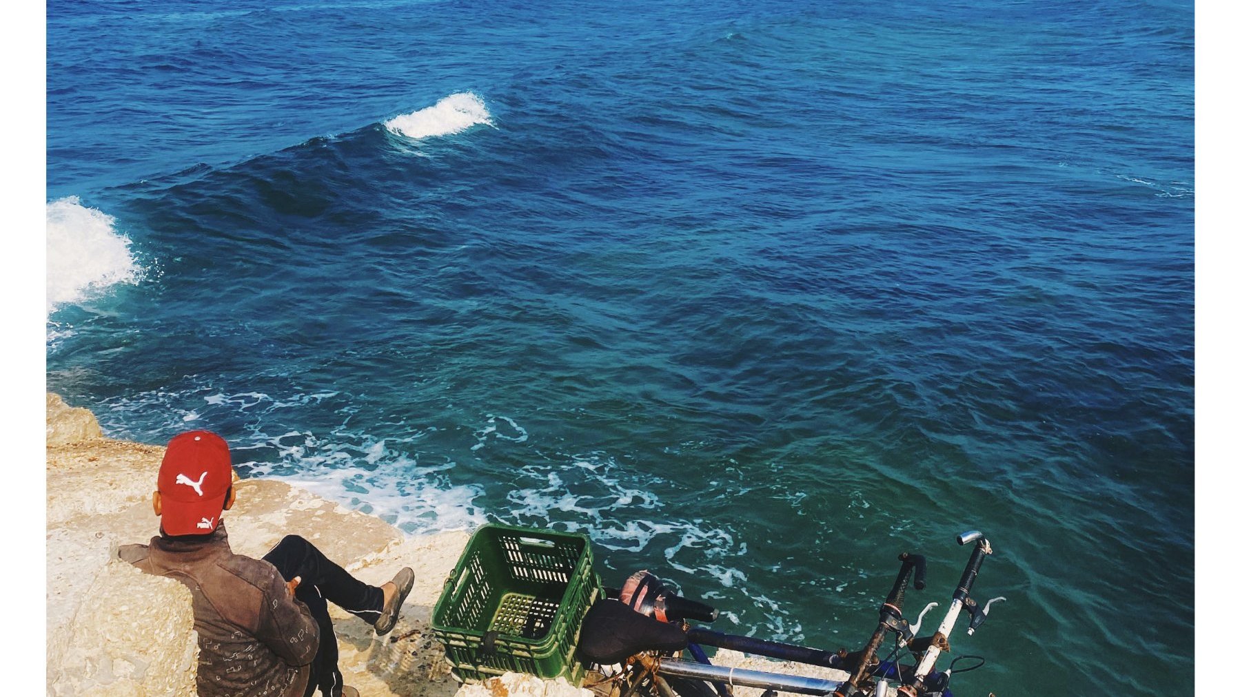 Un homme assis sur des rochers, face à une mer calme et bleue sous un ciel dégagé.