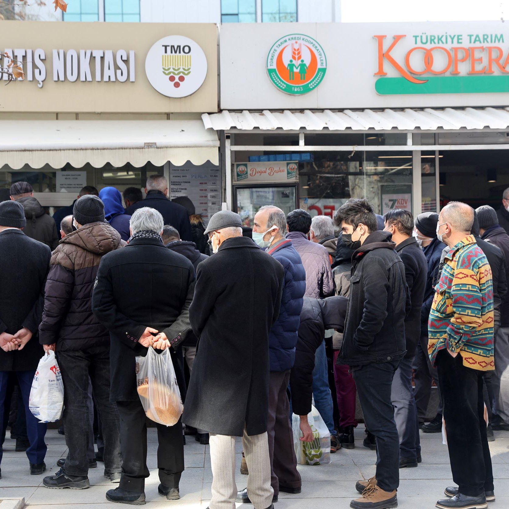 الصورة تظهر مجموعة من الأشخاص الواقفين في طابور أمام متجر يُظهر لافتة تدل على أنه نقطة بيع للمنتجات. يبدو أن معظم الأشخاص يرتدون ملابس شتوية وأنهم يحملون أكياسًا. الجو يبدو هادئًا، مع وجود أشجار حول المكان، بينما يبدو أن بعضها قد فقد أوراقه بسبب البرد. المتجر يحمل اسم "كوoperative" ويبدو أنه يبيع مواد غذائية أو منتجات محلية.