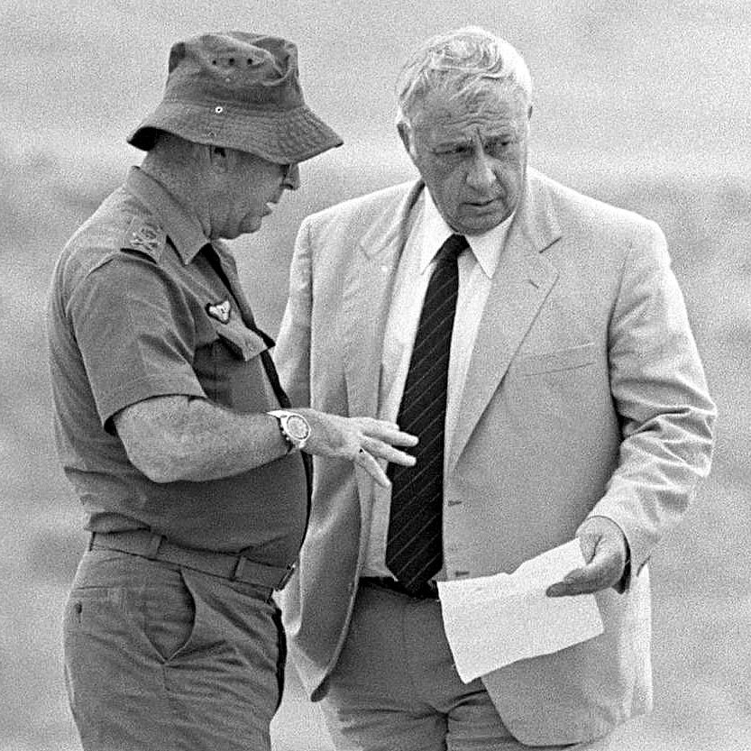 L'image montre deux hommes en train de discuter. L'un d'eux porte une tenue militaire, incluant un chapeau, tandis que l'autre est vêtu d'un costume clair. Ils semblent examiner un document ensemble et échanger des informations de manière sérieuse. L'arrière-plan est flou, suggérant un environnement extérieur. La scène dégage une impression de concentration et d'importance.