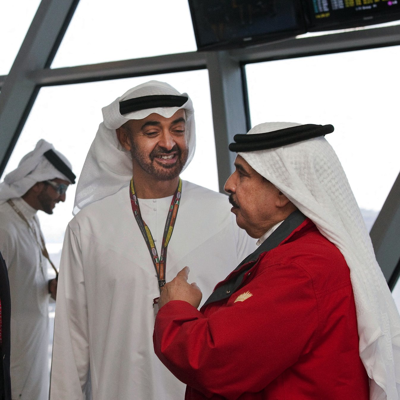 در این تصویر، سه مرد در یک مکان رسمی ایستاده‌اند. یکی از آن‌ها که لباس رسمی پوشیده، به نظر می‌رسد که از چهره‌ای جدی برخوردار است. دو نفر دیگر لباس سنتی عربی به تن دارند. یکی از آن‌ها لباسی سفید و دیگری لباسی قرمز به همراه کلاه عربی دارد. آن‌ها به طور دوستانه با یکدیگر صحبت می‌کنند و در پس‌زمینه می‌توان تعدادی دیگر از افراد را دید که در حال حرکت هستند. فضای تصویر نشان‌دهنده یک مکان شلوغ و مدرن است.