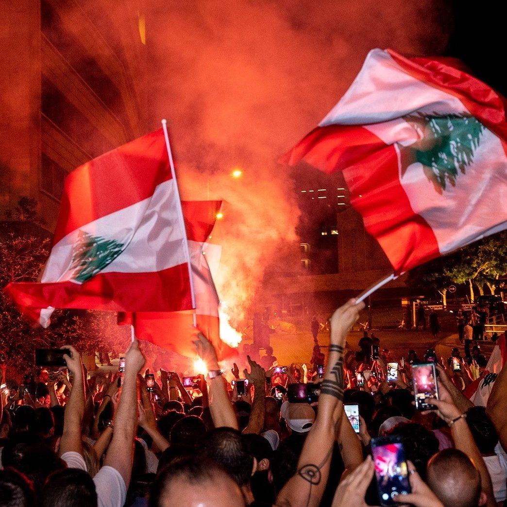 تظهر الصورة تجمعًا حاشدًا في أحد الشوارع، حيث يحمل المتظاهرون أعلام لبنان. تضيء الأجواء بألوان النيران والدخان، مما يخلق جوًا احتفاليًا أو تعبيريًا. يُلاحظ أن الكثير من الأشخاص يستخدمون هواتفهم لتسجيل اللحظة أو التقاط الصور، بالإضافة إلى الأضواء الساطعة التي تضيف حيوية للمشهد.